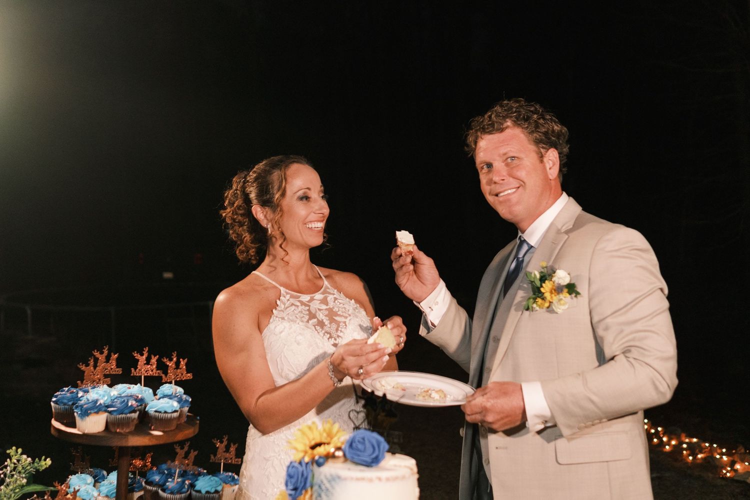 Smiling bride and groom share wedding cake outdoors at night with decorated dessert table behind them.