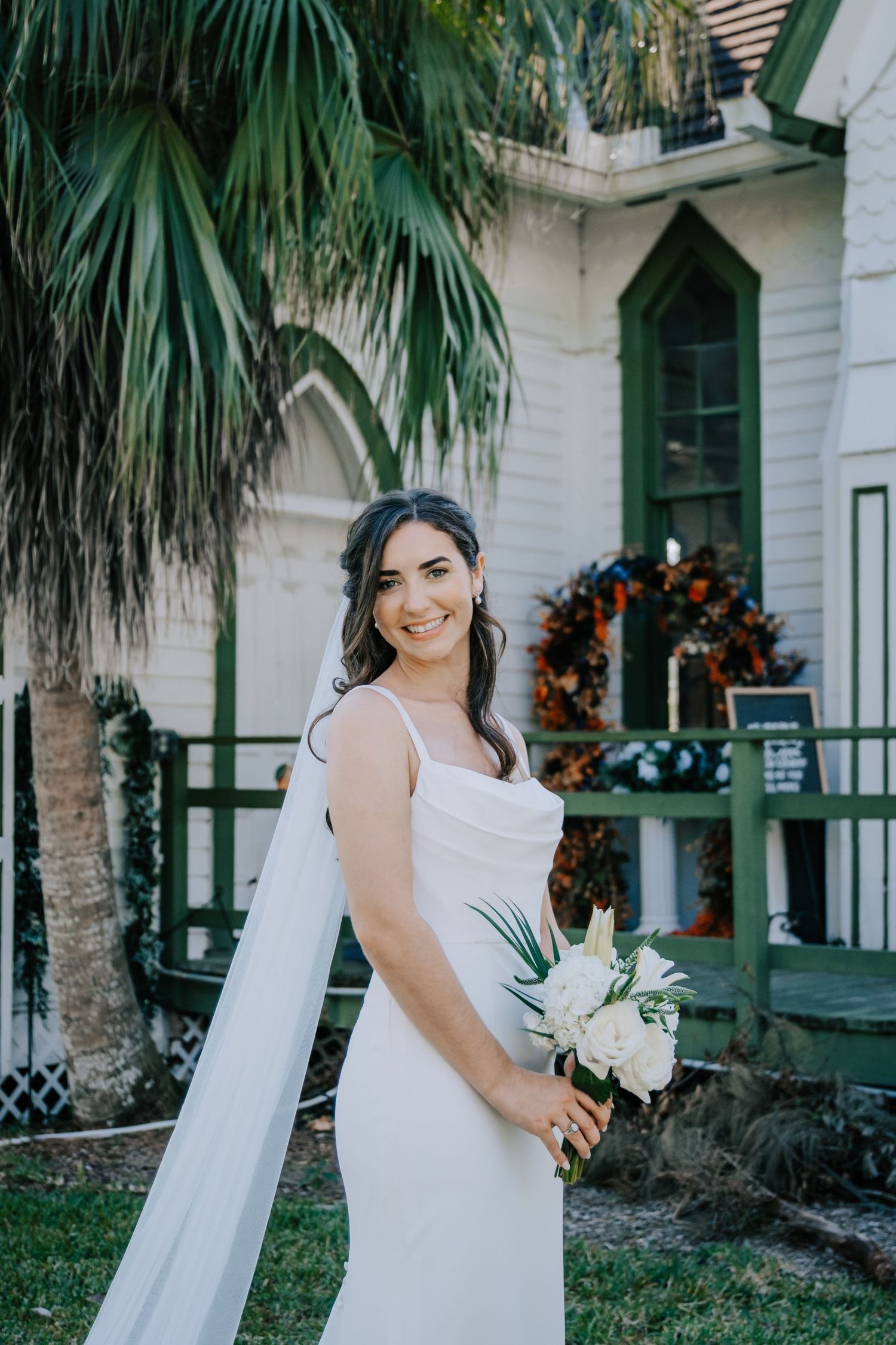 A bride in a white dress poses with her bouquet in front of a historic building with palm trees.