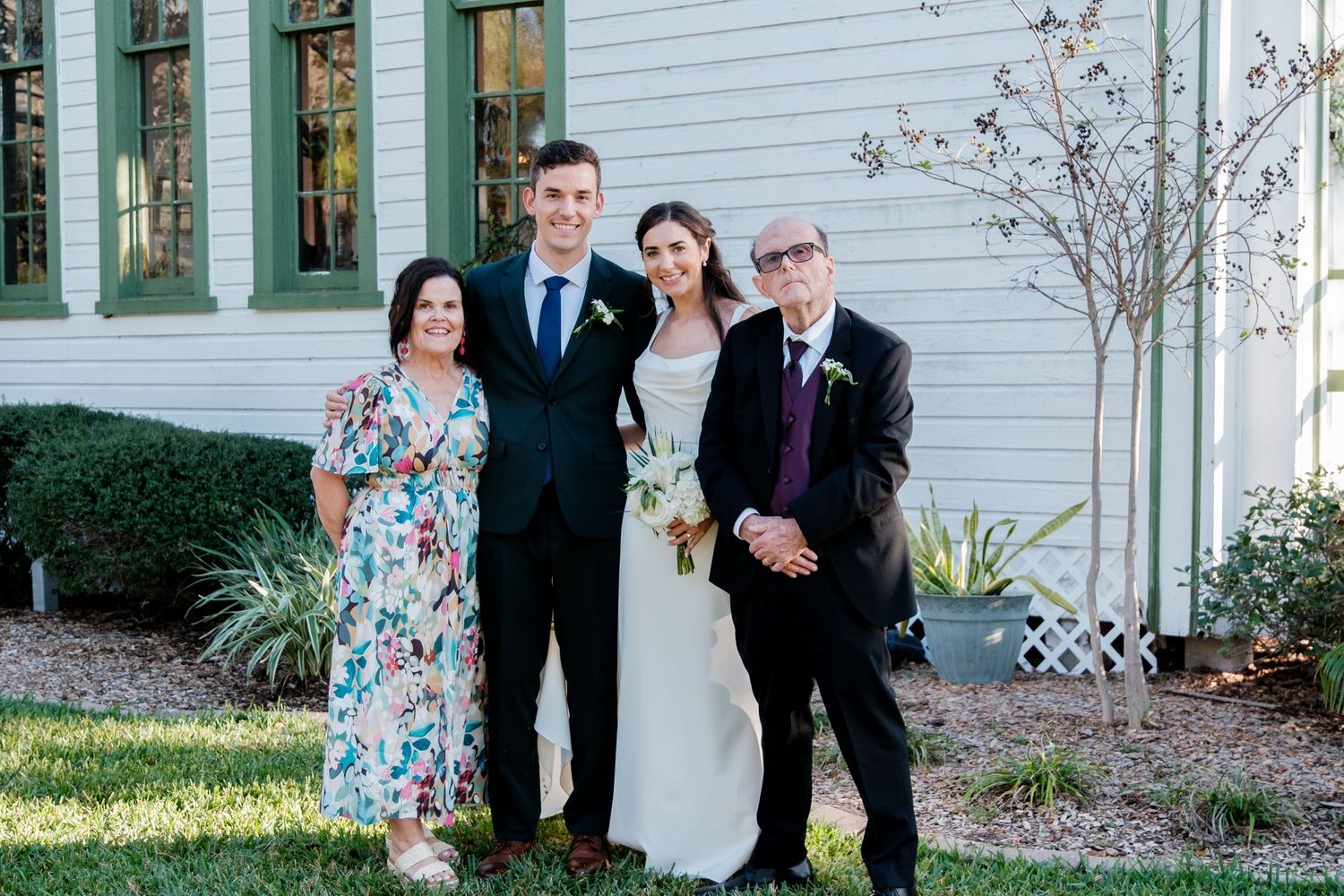 Wedding party members gather outside a white church building with green trim.