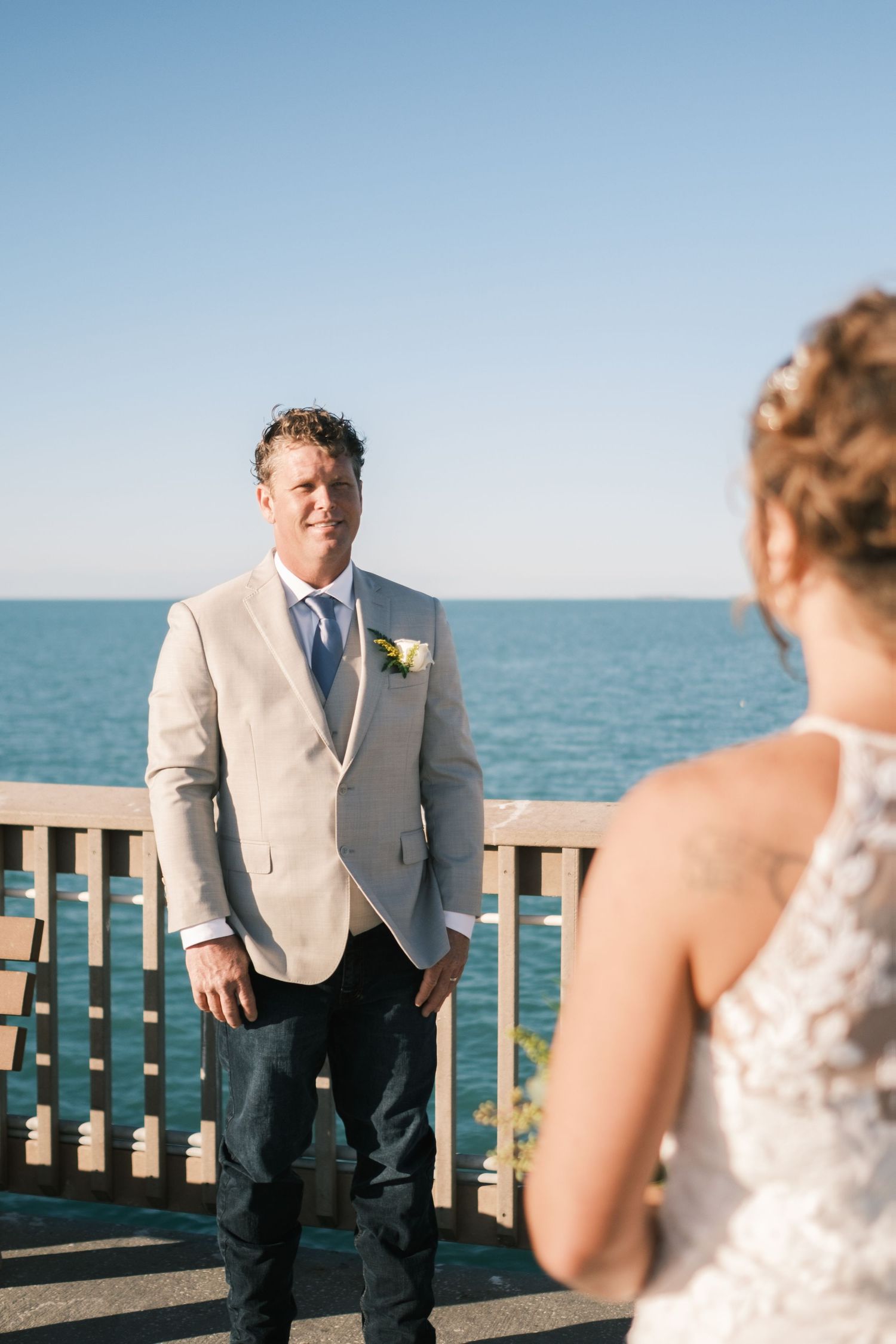 Groom smiling at bride during oceanside wedding ceremony with blue sea and clear sky backdrop.
