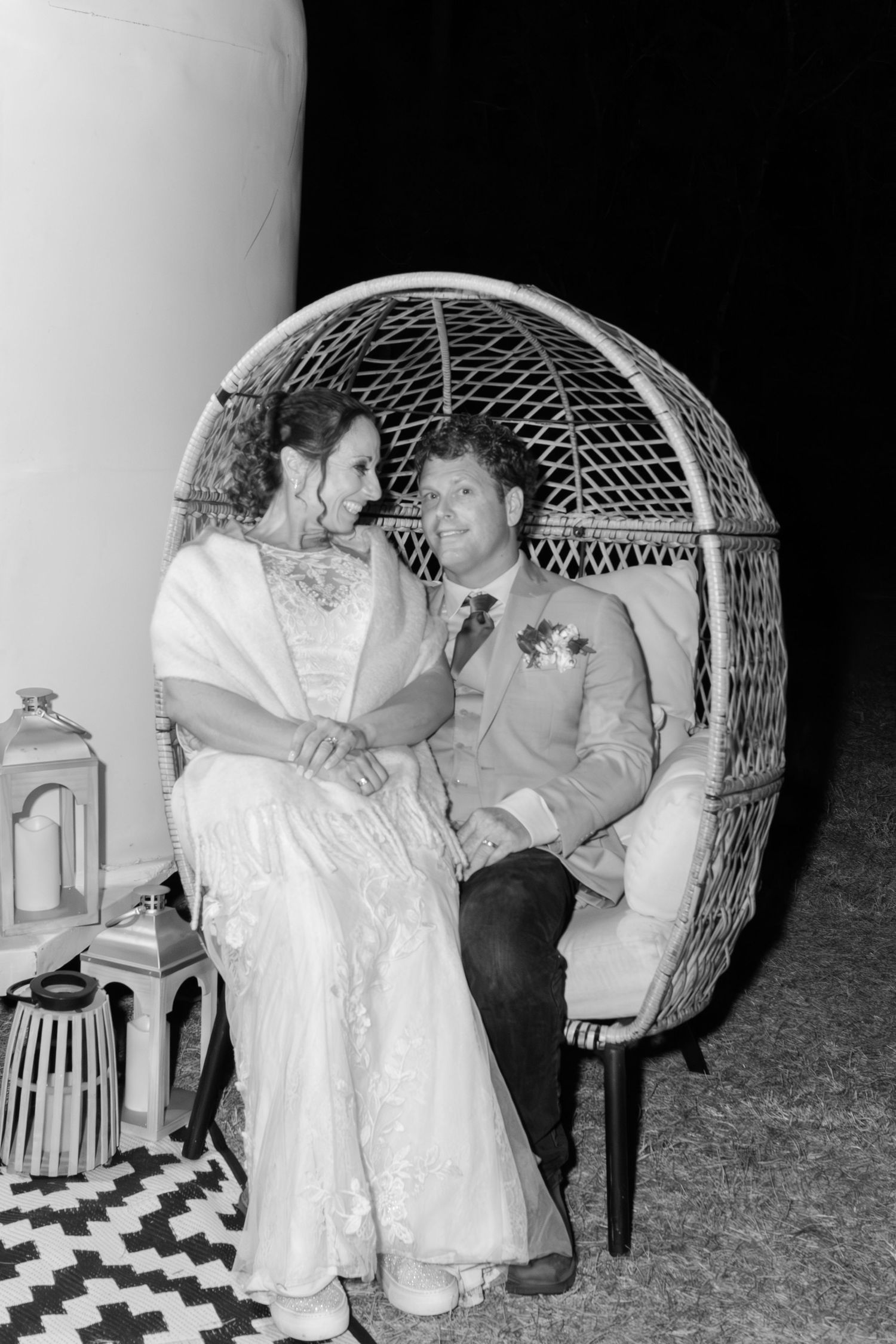 Bride and groom sit together in wicker egg chair sharing intimate moment at nighttime reception.