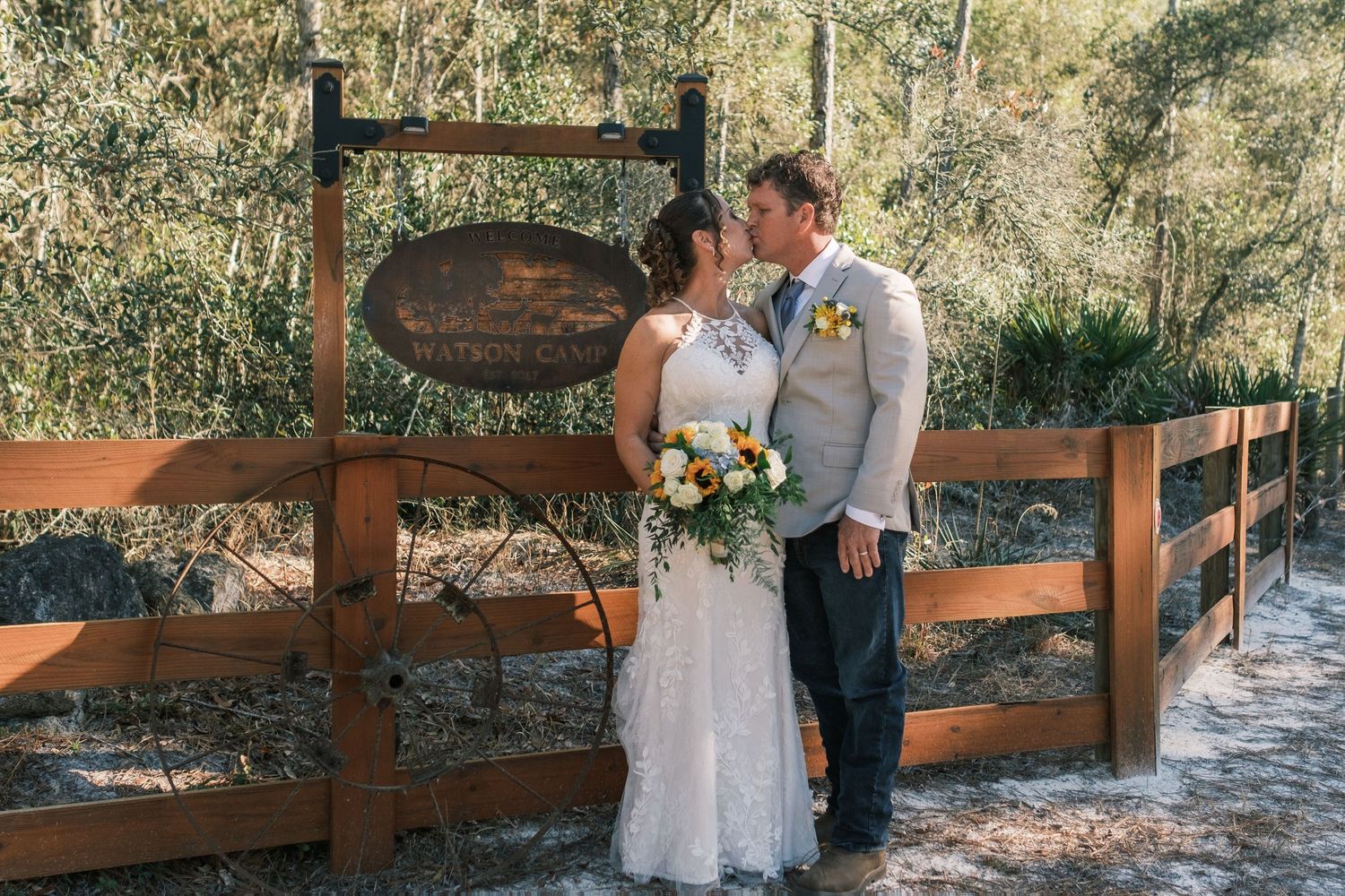 Bride and groom share a kiss beside a rustic wooden fence sign surrounded by lush green trees.