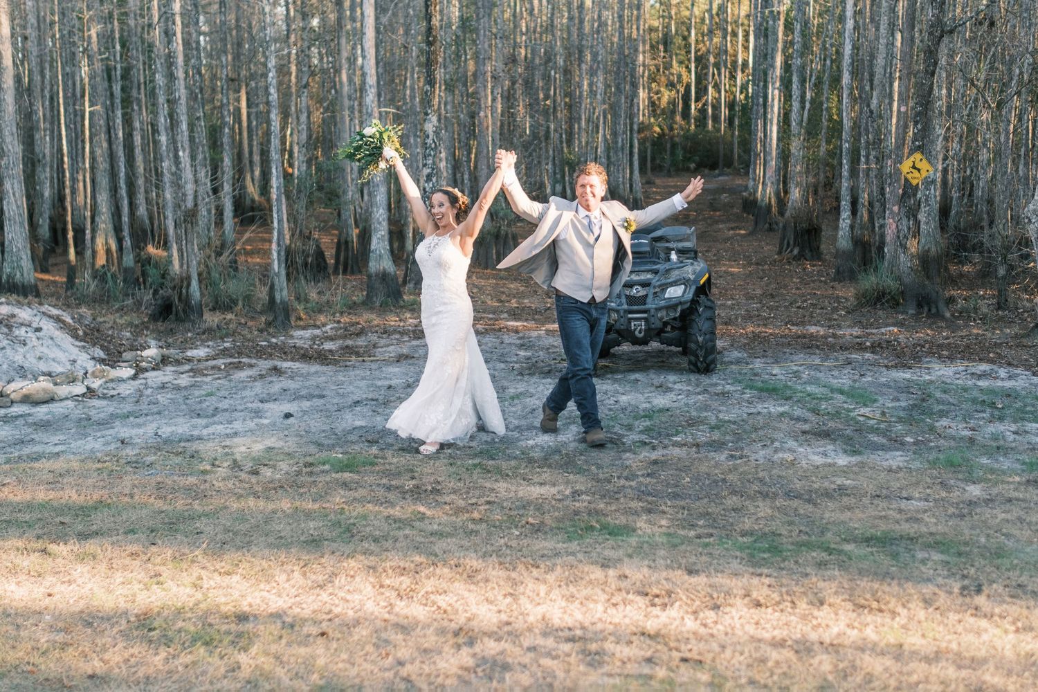 Joyful newlyweds celebrate with raised arms near an ATV in a rustic woodland setting.