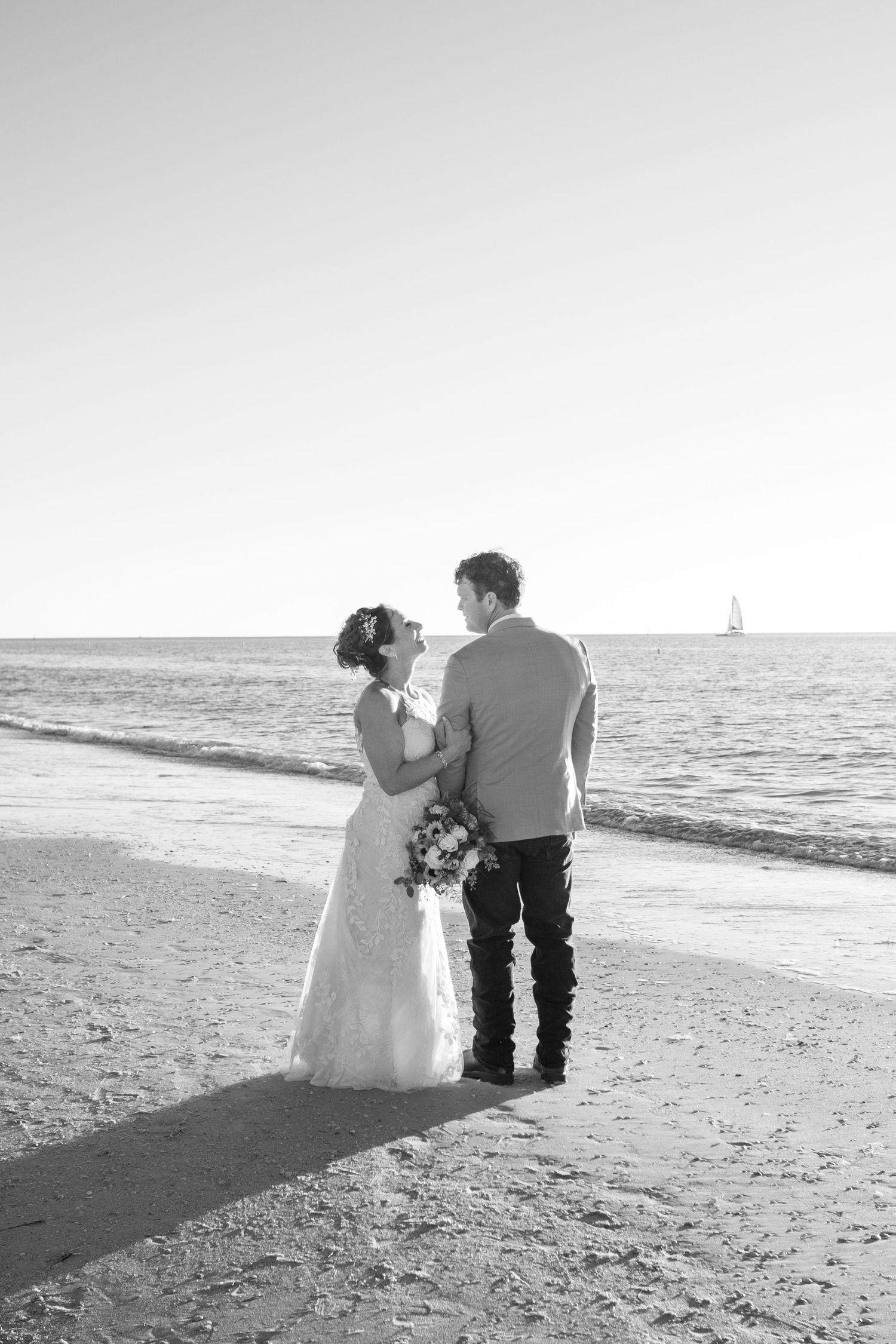 Black and white photo of bride and groom embracing on beach with sailboat in background.