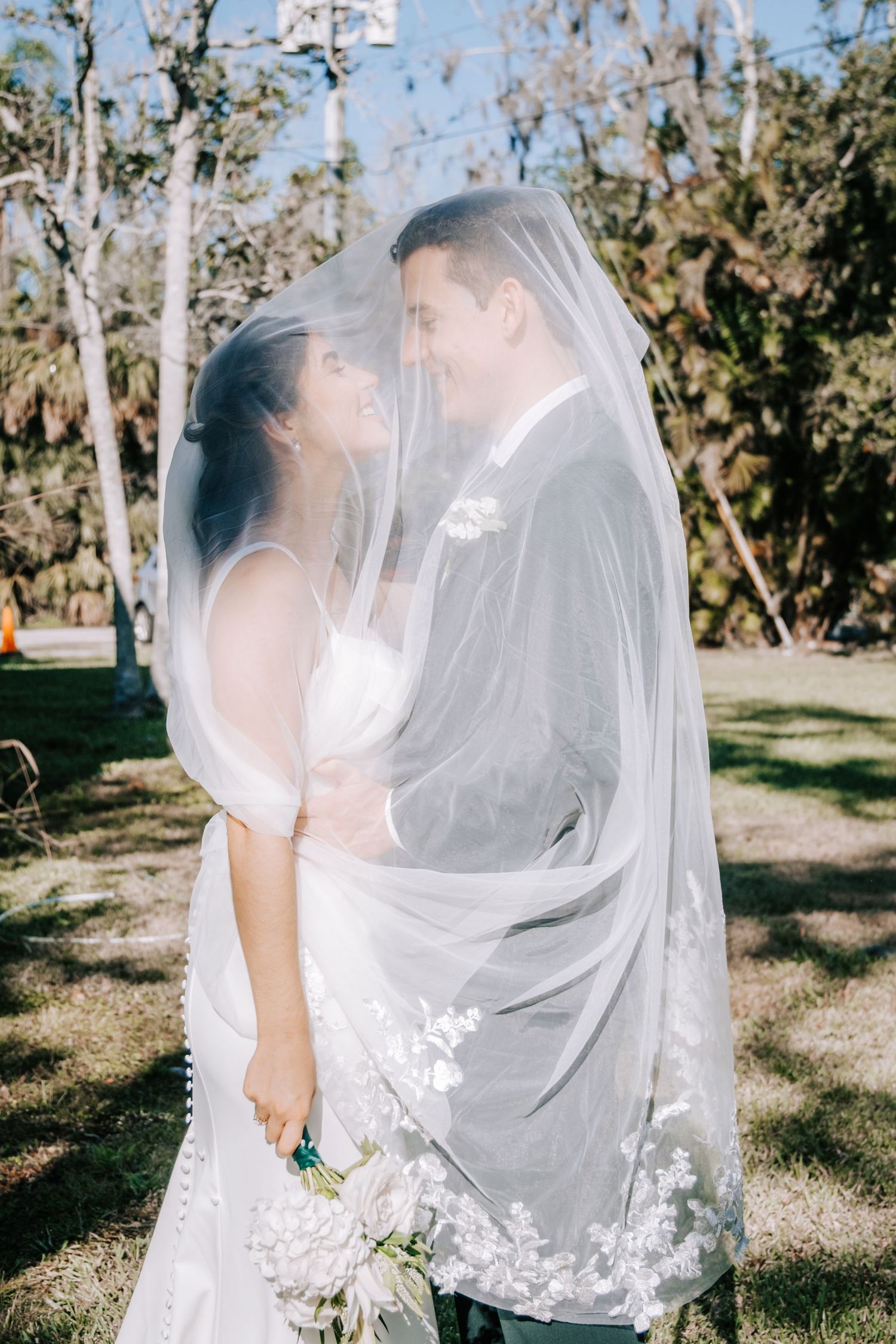A tender moment between newlyweds under a delicate veil in a garden setting.