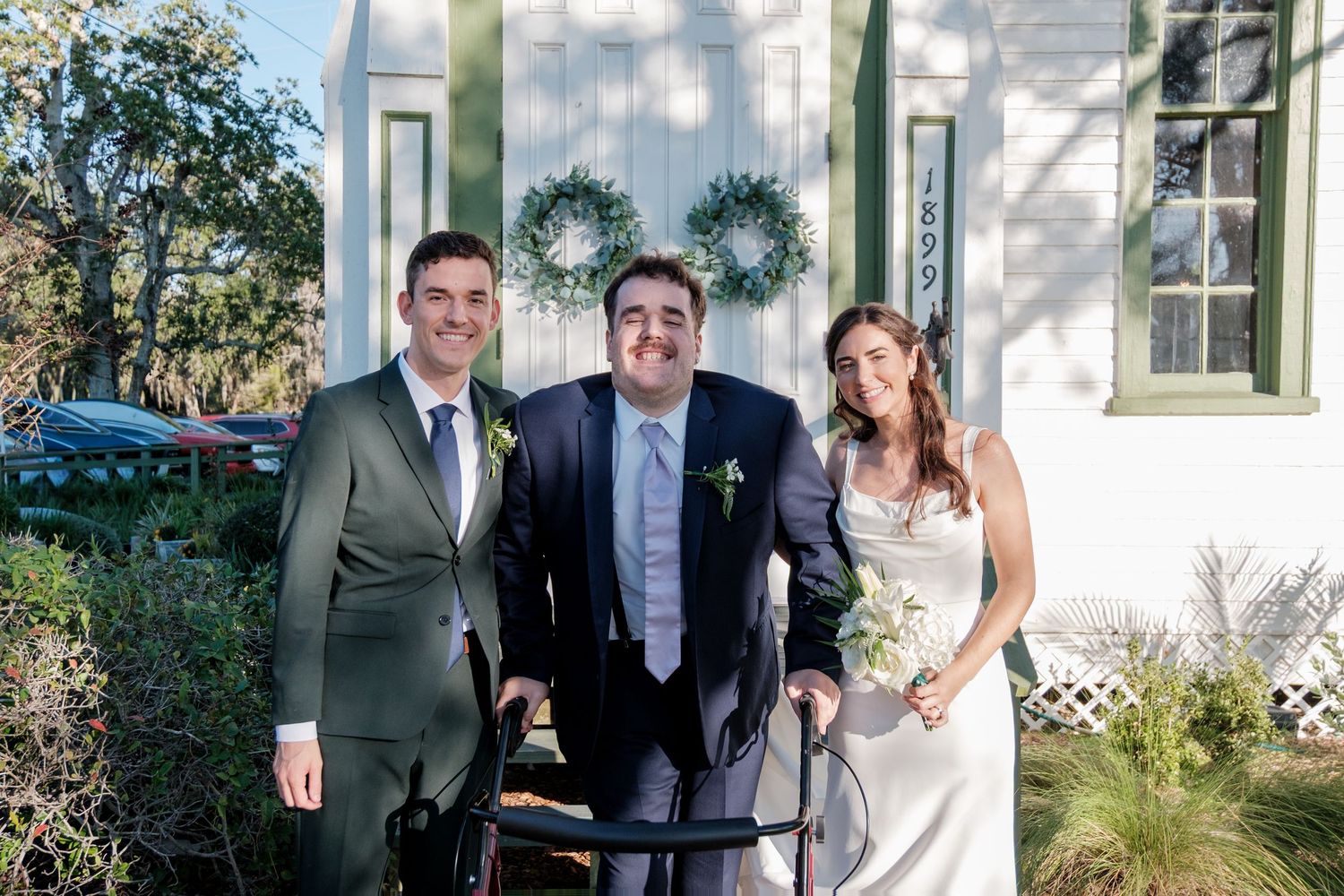 A wedding party poses for a photo outside a white chapel with green trim.