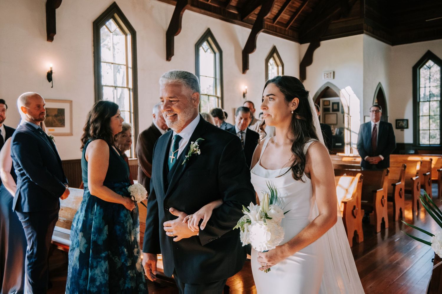 Wedding party members in navy and formal attire walk through a sunlit church hallway.
