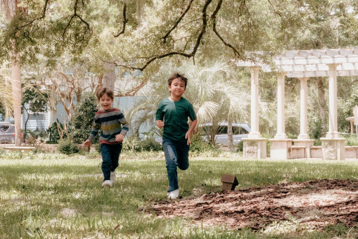 Two children run and play together in a park with picnic tables and trees on a sunny day.