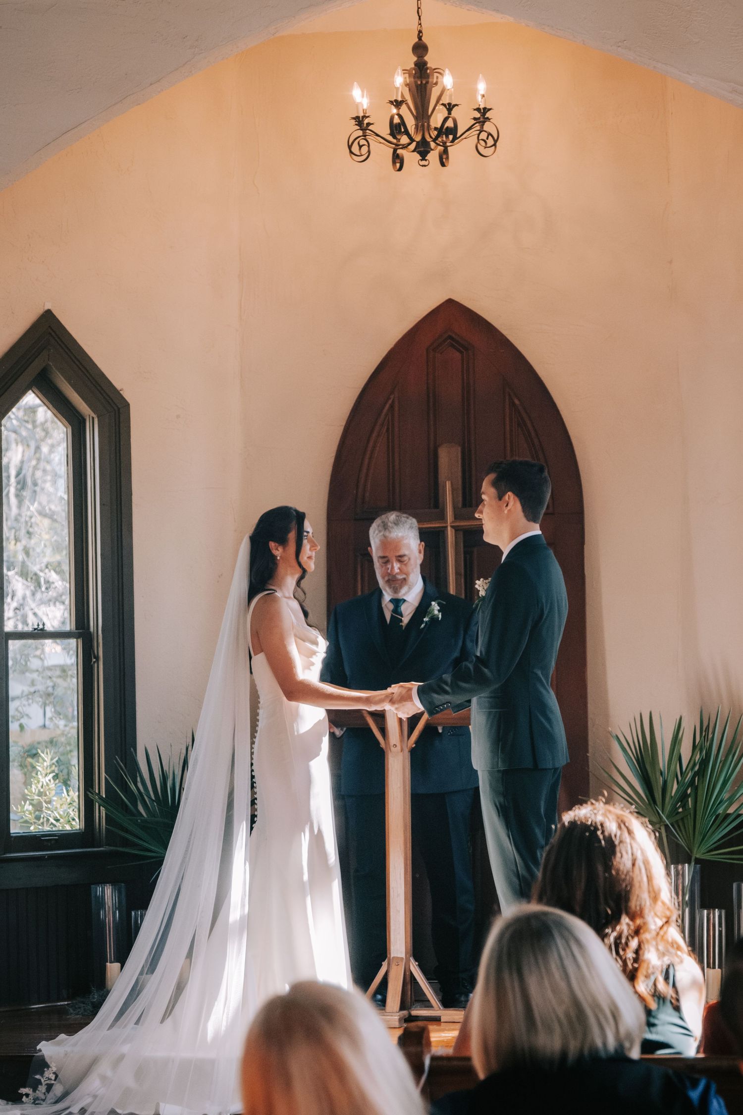 A wedding ceremony takes place in a rustic chapel with arched windows and indoor plants.