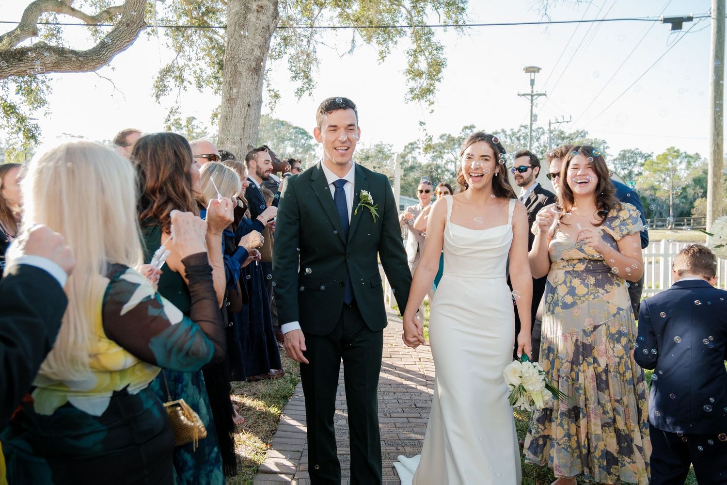 Newlyweds walk down aisle while guests celebrate at outdoor ceremony under oak trees.