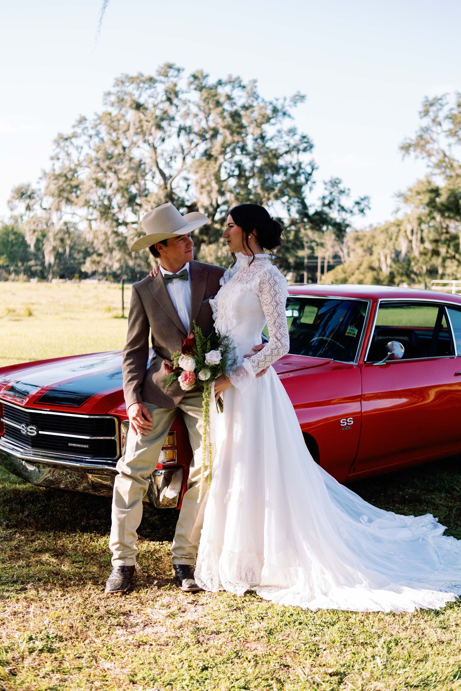 Bride and groom pose by a red Chevrolet SS muscle car outdoors, groom in cowboy hat holding roses.