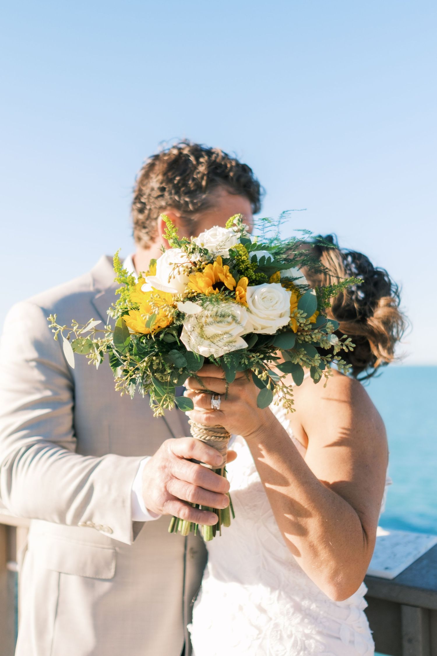 Bride and groom kiss behind a lush bouquet of white roses and yellow flowers with ocean in background.