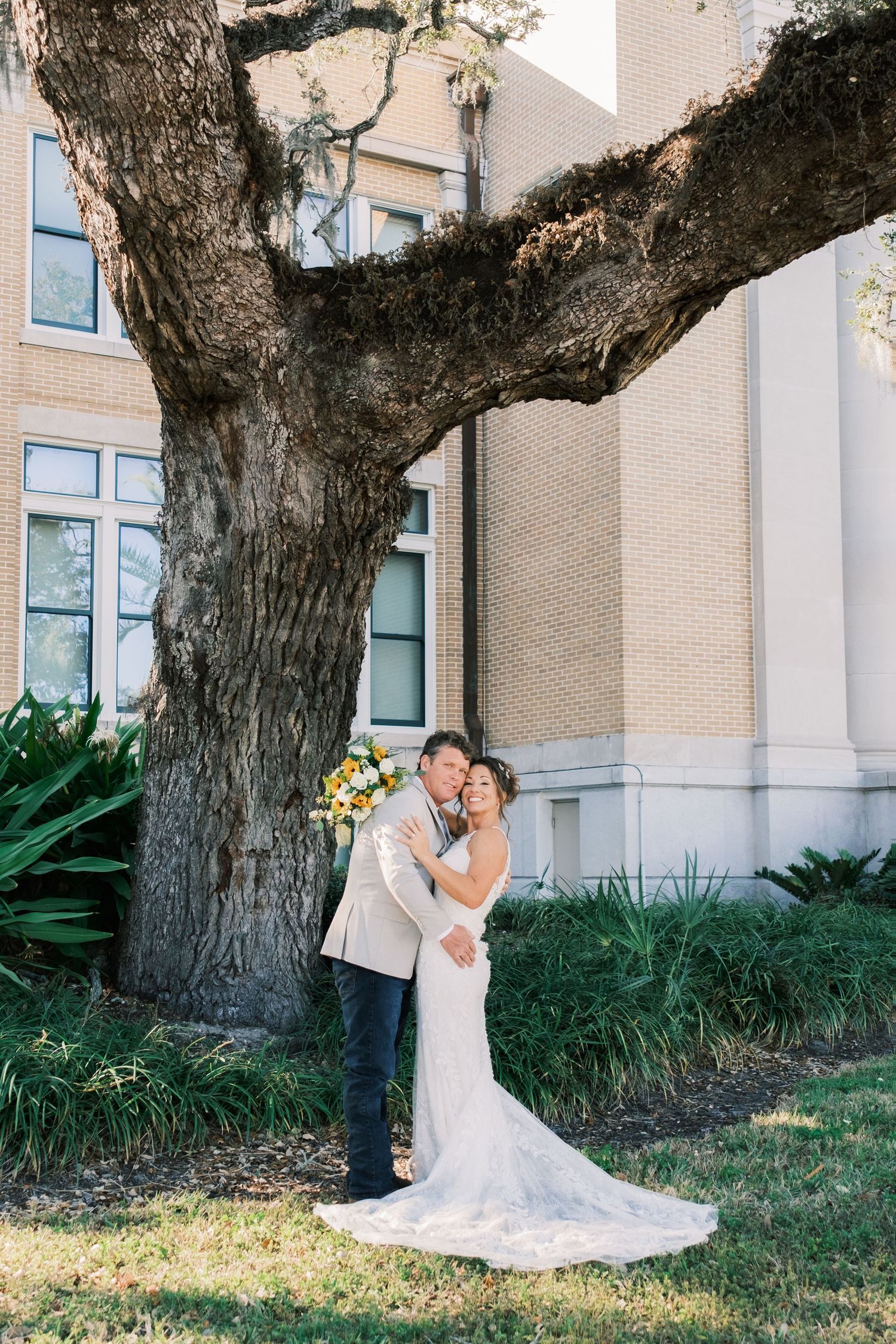 Bride and groom embrace under a large oak tree outside a brick building, bride holding sunflower bouquet.