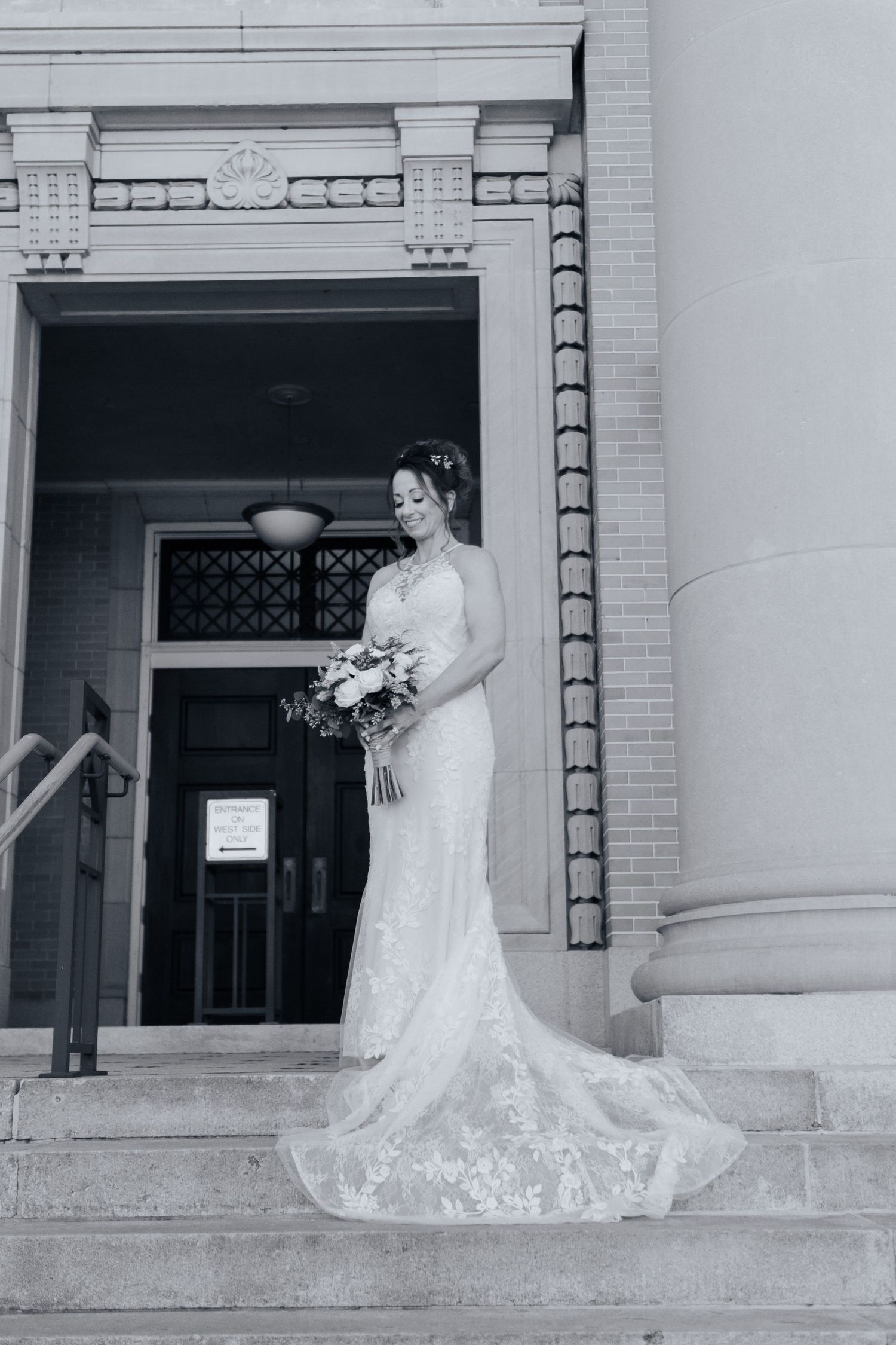 Black and white photo of bride in lace wedding gown holding bouquet on steps of a classic building.