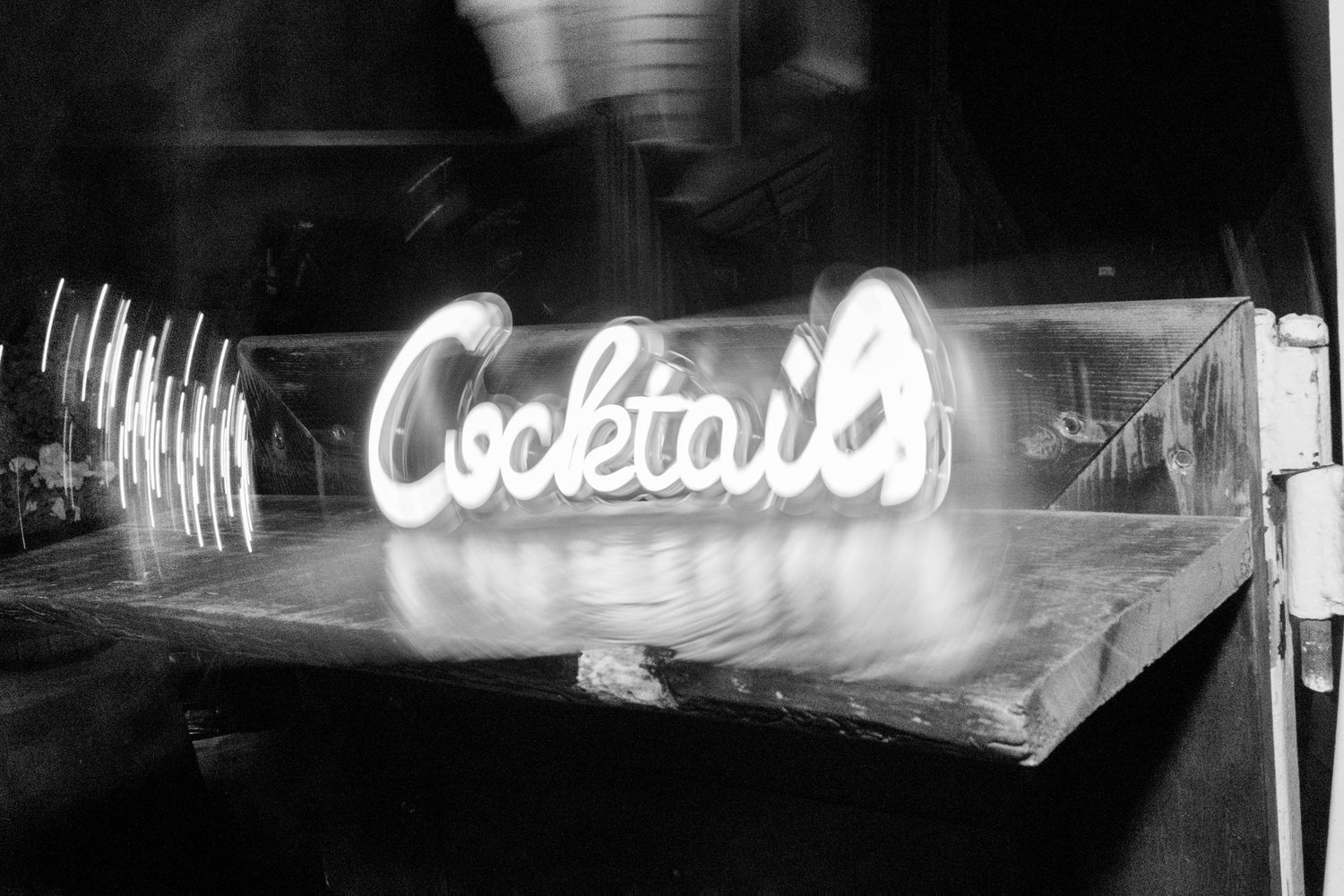 Glowing neon Cocktails sign on a bar counter photographed in black and white at a wedding reception.
