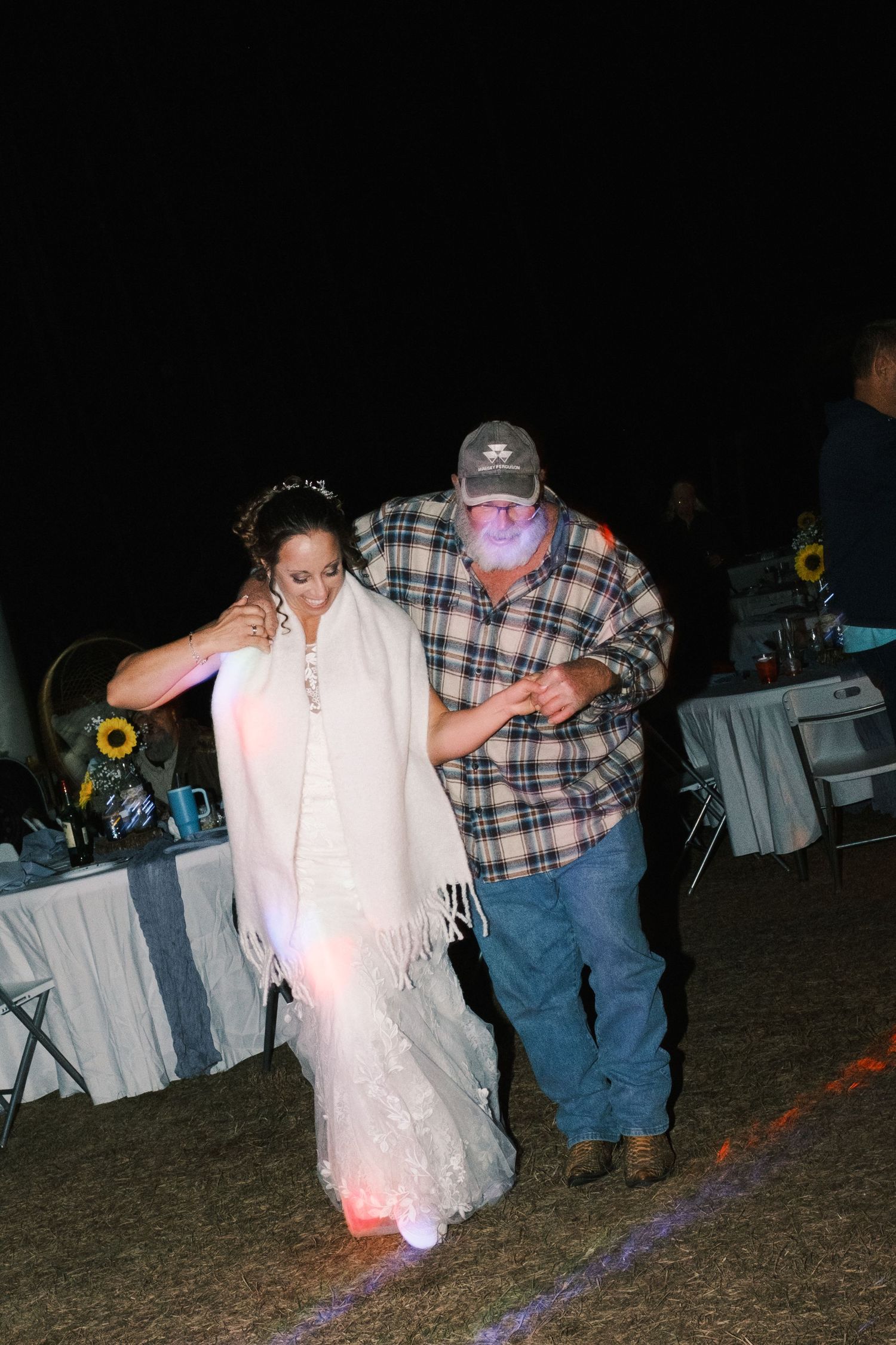 Bride in white lace gown and shawl dancing with older man in plaid shirt at outdoor wedding reception.