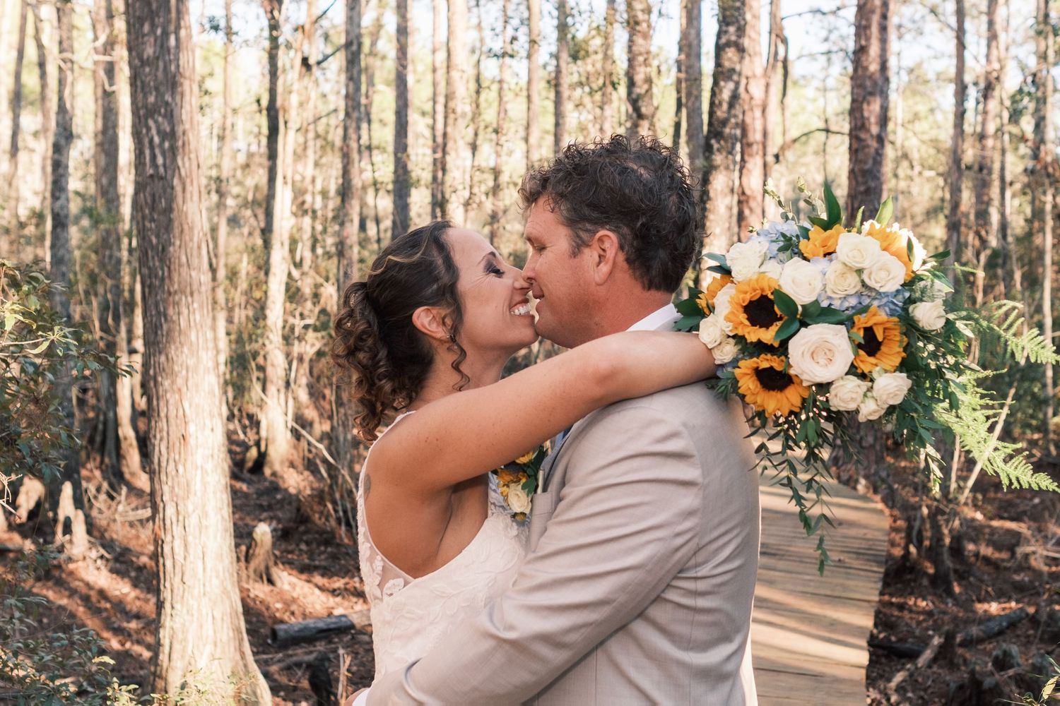 Bride and groom share a romantic kiss in a sunlit forest, bride holding a sunflower and rose bouquet.