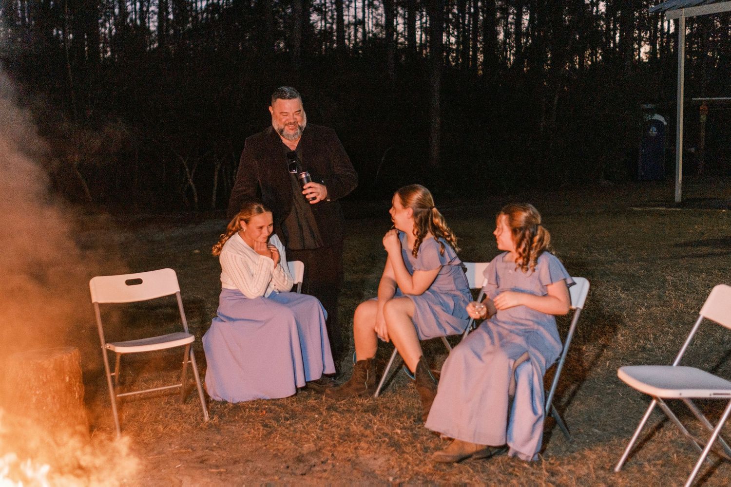 Man socializes with three bridesmaids in blue dresses seated around a glowing campfire at night.