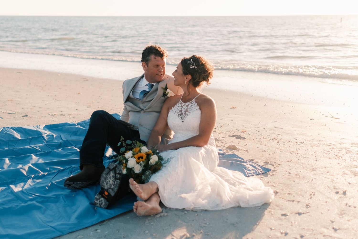 Bride and groom sitting on blue blanket on beach at sunset, bride holding colorful bouquet.