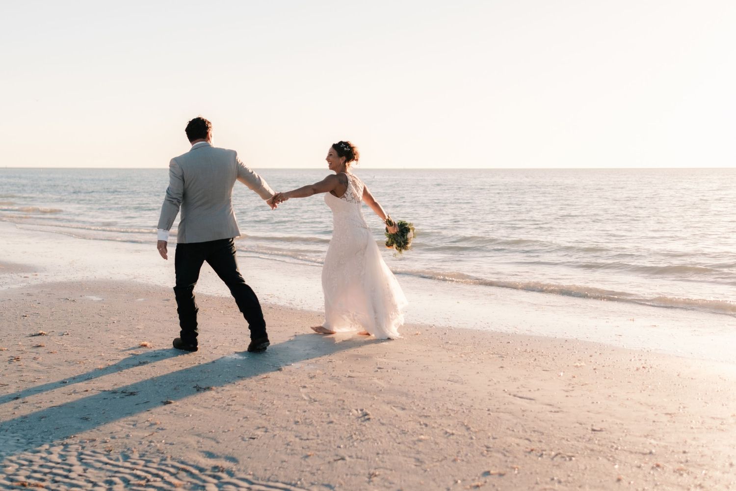 Bride and groom holding hands walking along sandy beach shoreline at sunset.