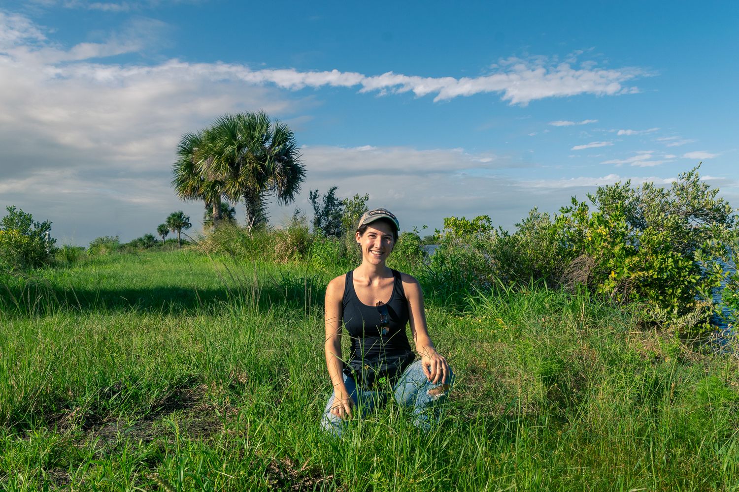 Woman kneeling in lush green field with palm trees and blue sky in natural Florida landscape setting.