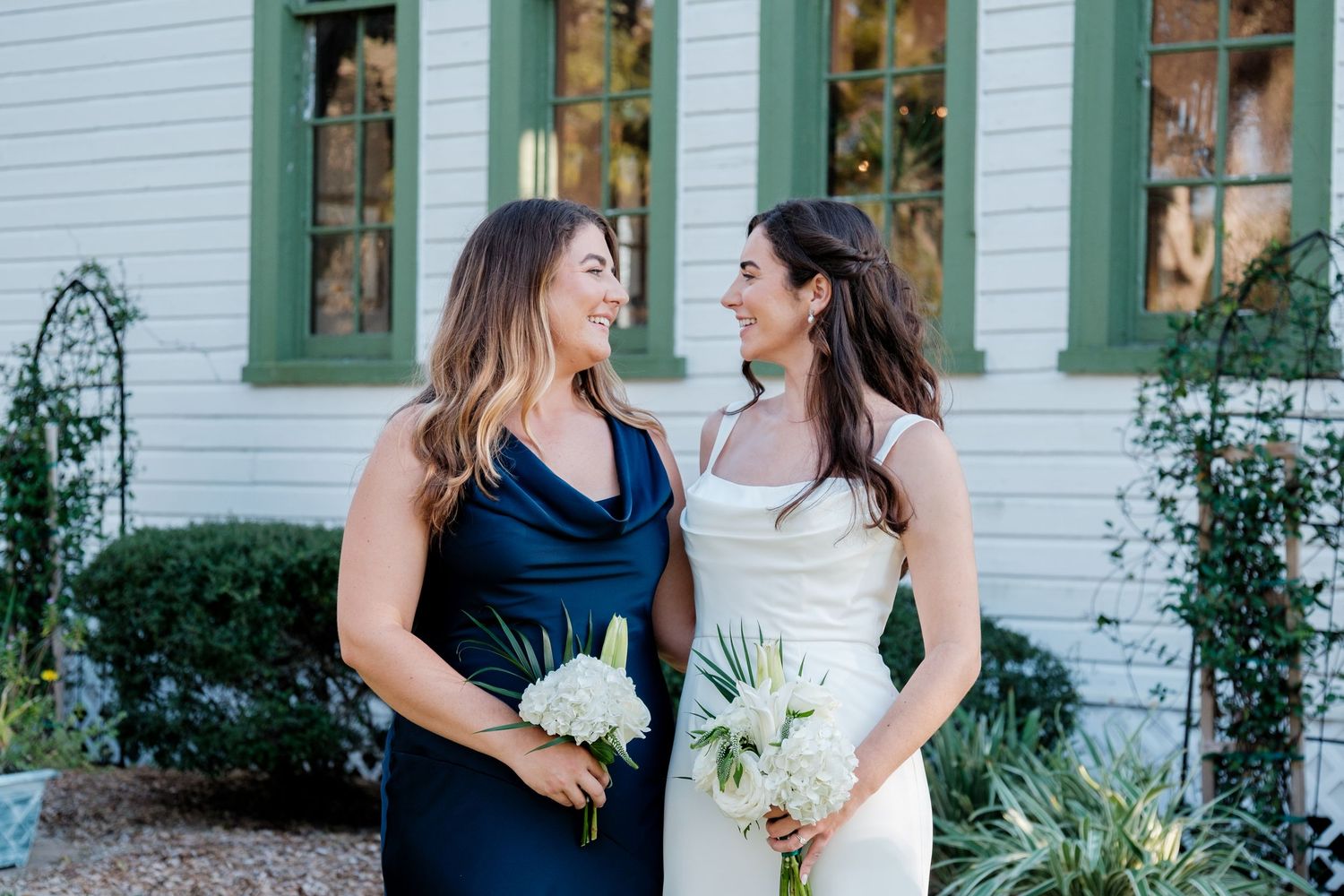 Two women in elegant dresses share a joyful moment while holding bouquets outside a white house.