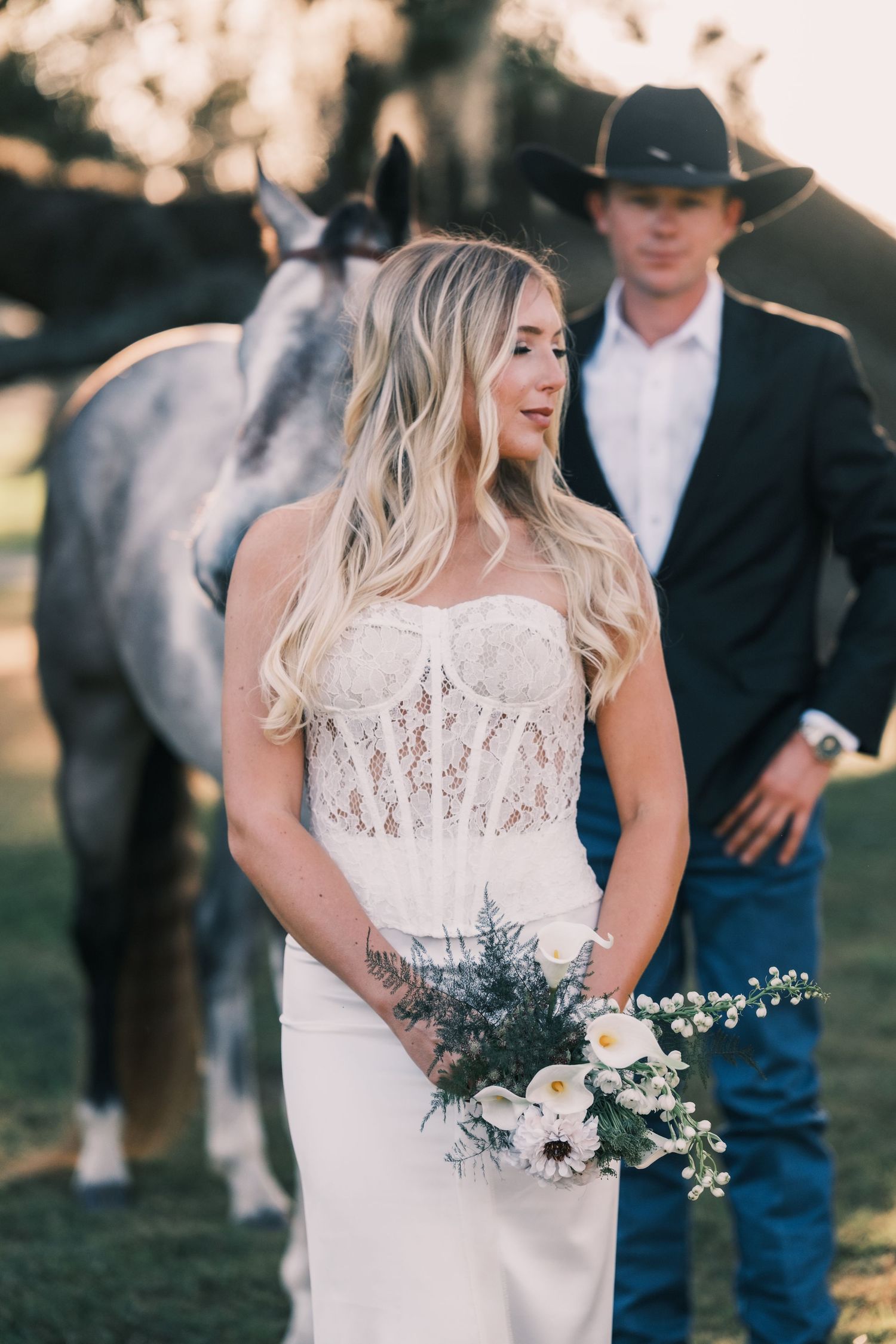 Rustic country wedding scene with couple in formal attire and white horse in background during golden hour.