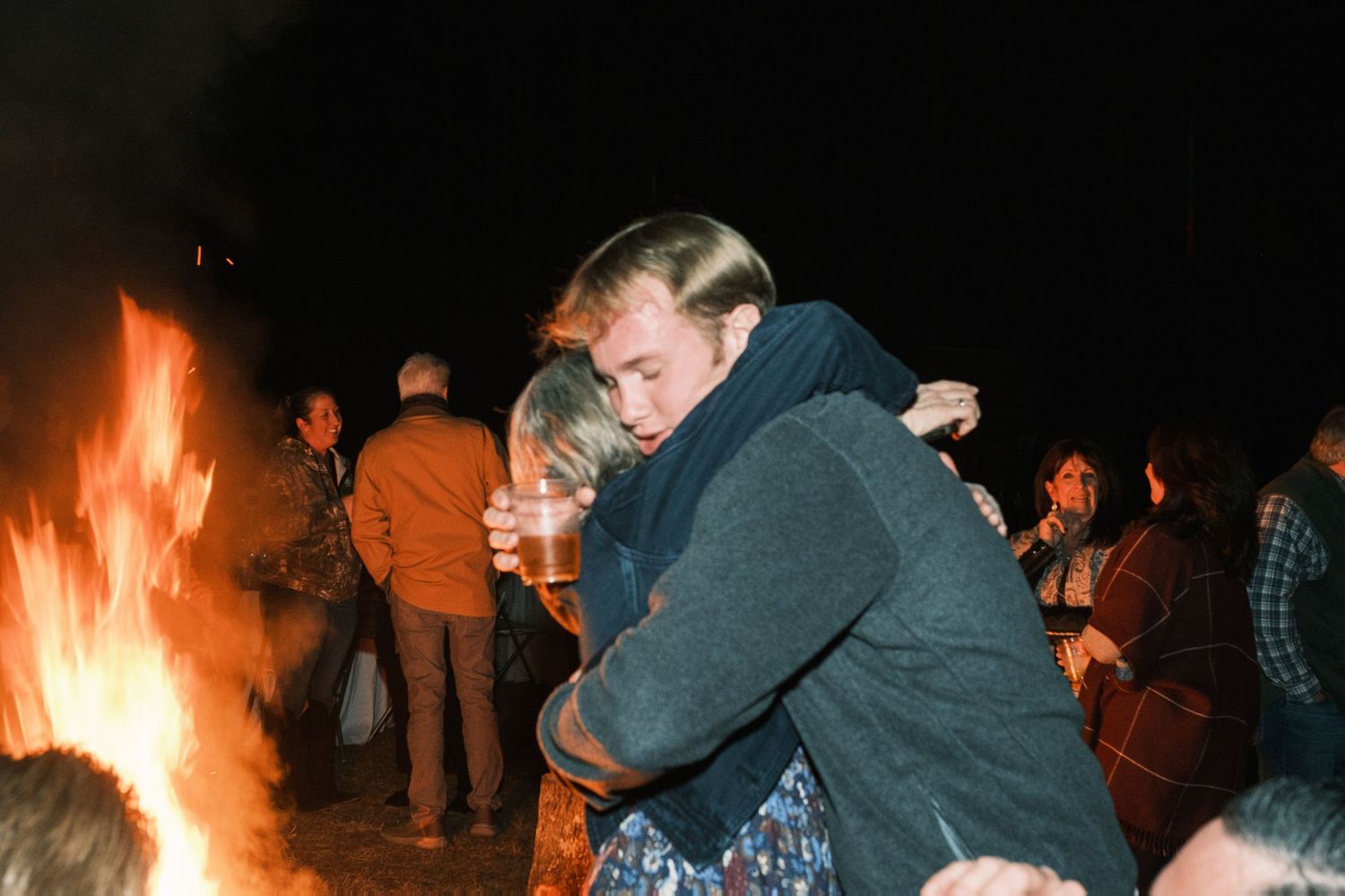 Two guests embracing near a bonfire at an outdoor nighttime wedding celebration.