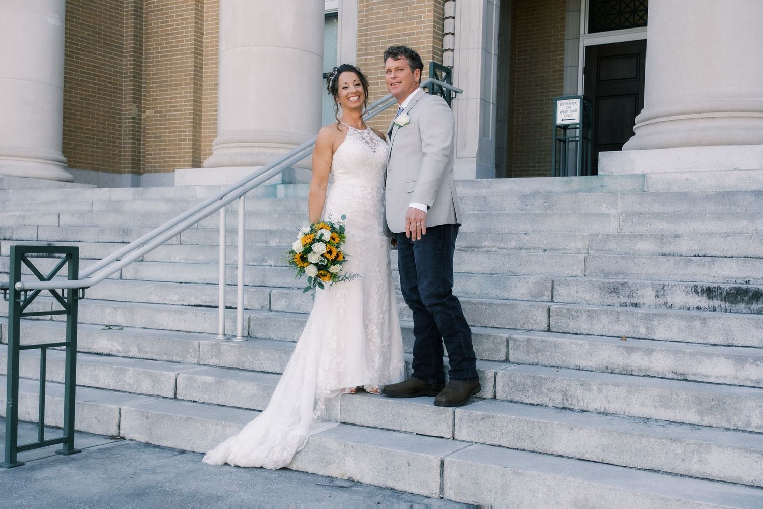 Bride holding colorful bouquet and groom in gray suit posing together on stone building steps.