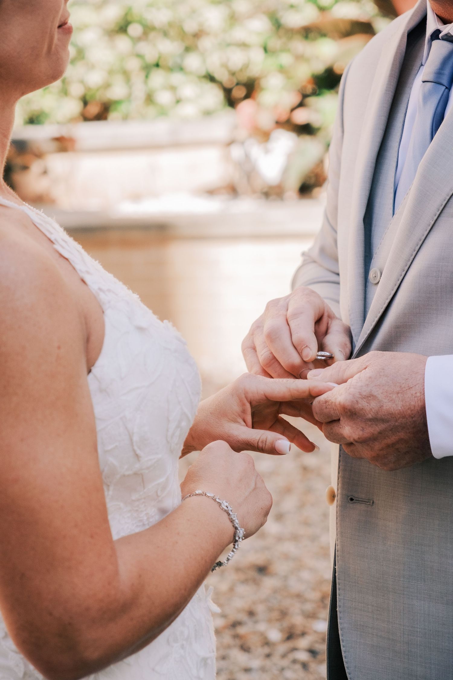 Groom placing a wedding ring on bride's finger during outdoor ceremony, bride wearing white lace dress and diamond bracelet.