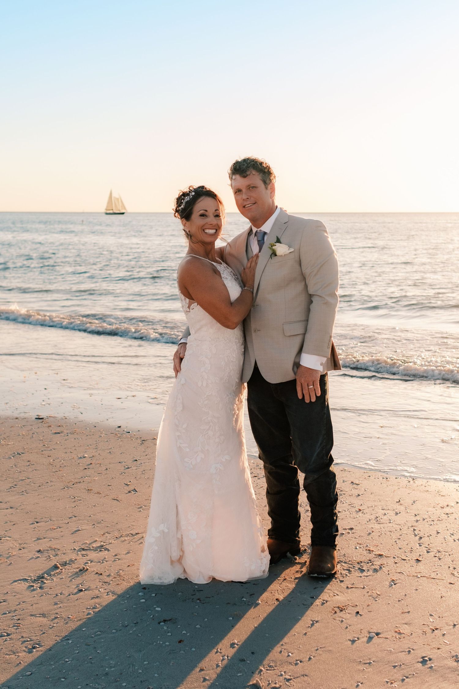 Bride and groom stand on a beach at golden hour, smiling, with a sailboat visible on the horizon.