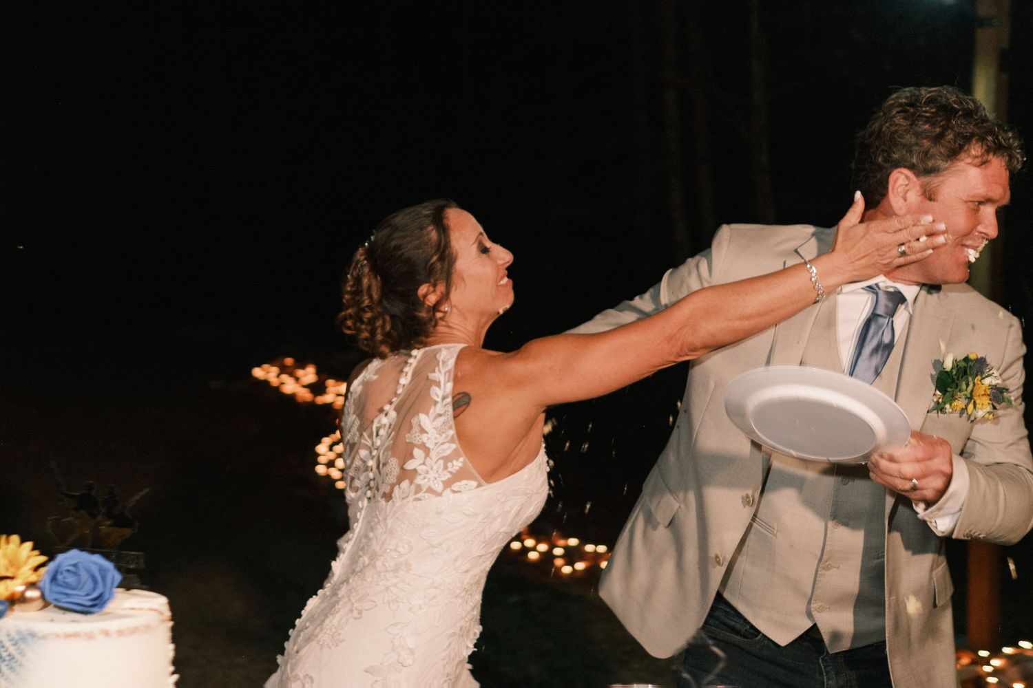 Bride playfully smashing wedding cake into groom's face during reception celebration at night.