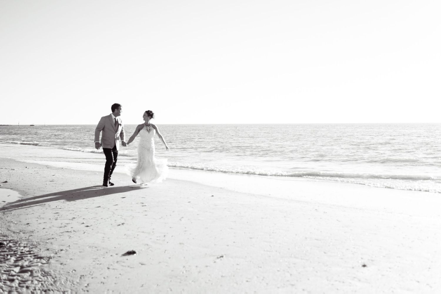 Black and white photo of bride and groom holding hands walking along a sandy beach shoreline.