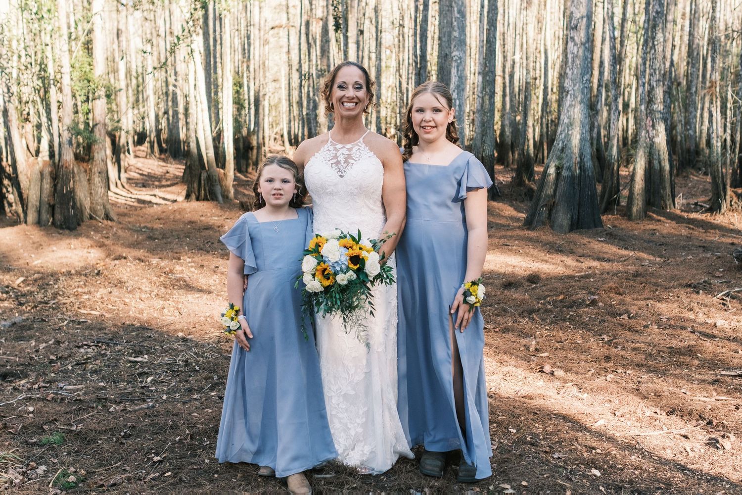 Bride in lace halter gown poses with two bridesmaids in blue dresses among tall trees.
