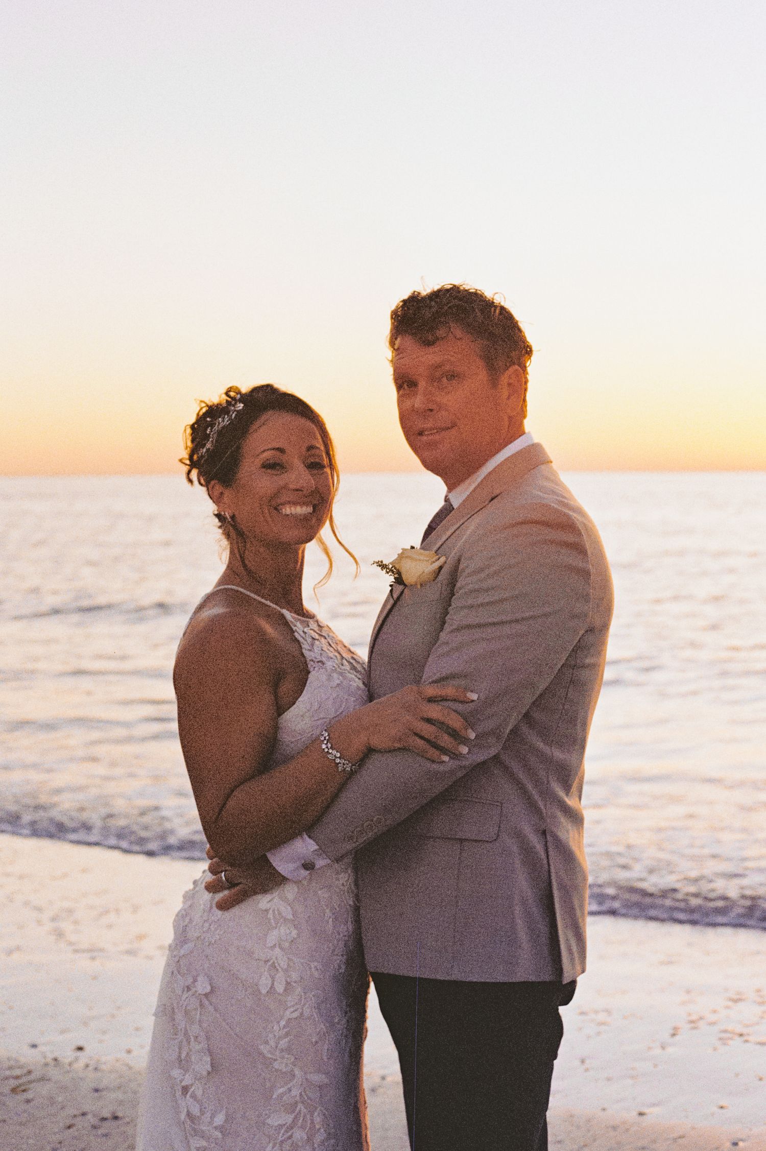 Bride and groom embrace on the beach at sunset with ocean waves behind them.