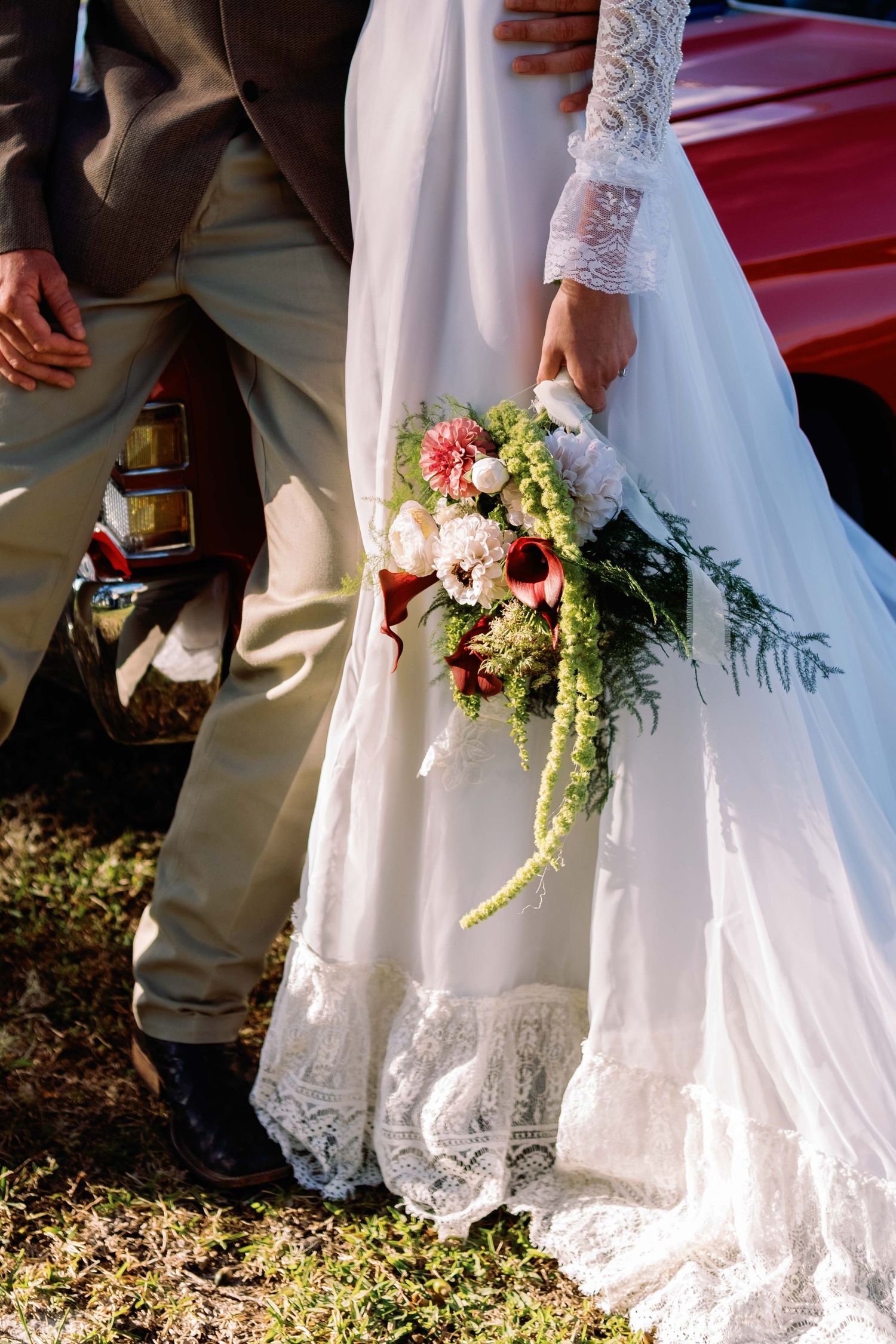 Bride in white lace wedding dress holding a cascading bouquet of red and pink flowers beside groom in gray suit.