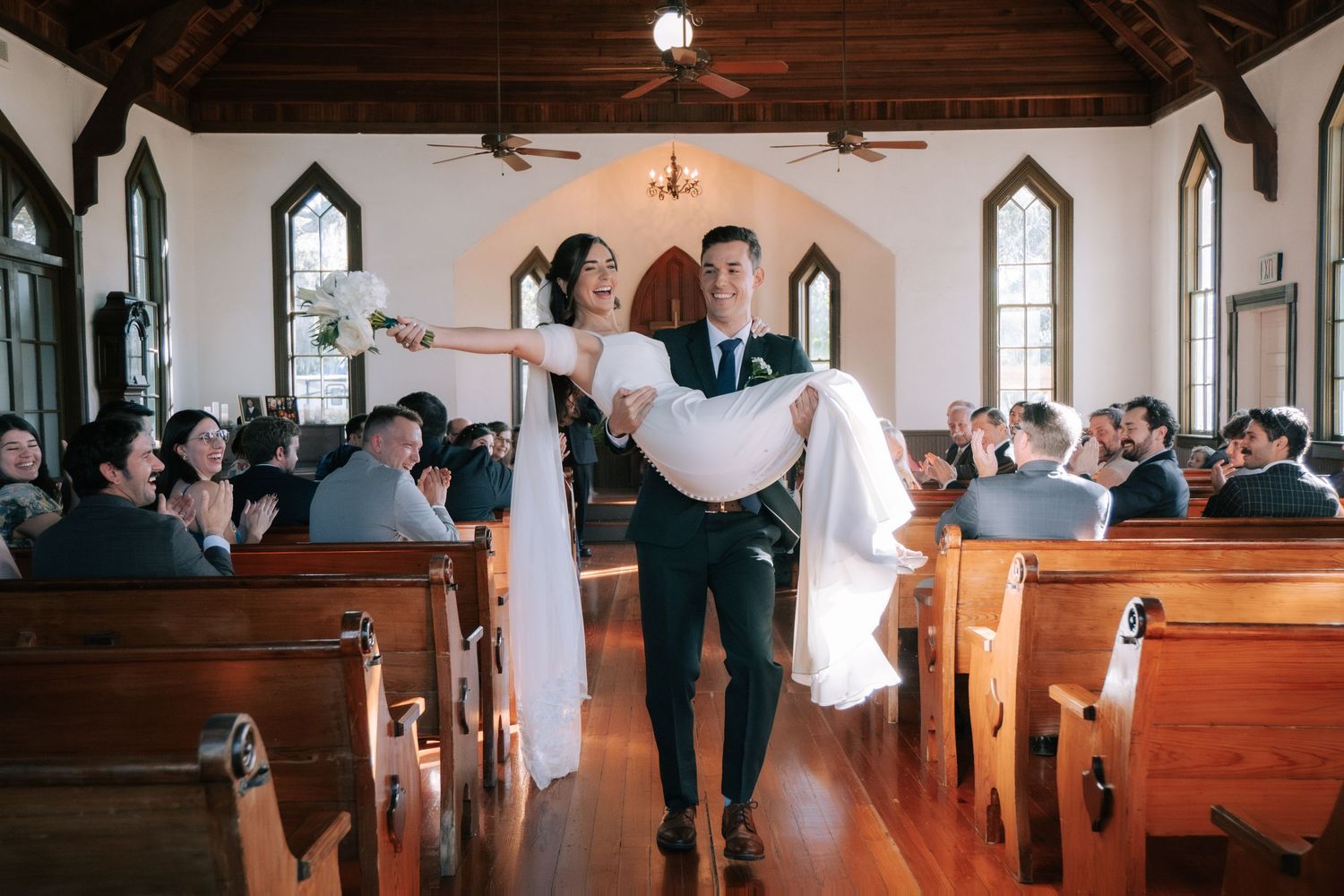 Groom carrying bride down the aisle of a charming white chapel with wooden pews and guests celebrating.