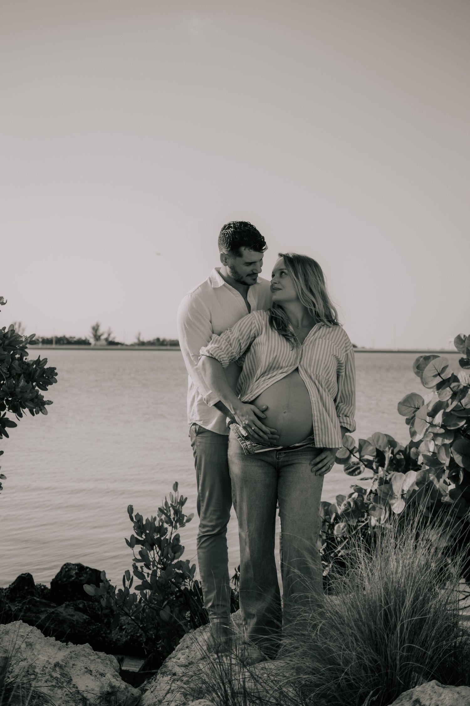 A romantic black and white maternity photo of a couple embracing by the water's edge with coastal vegetation.
