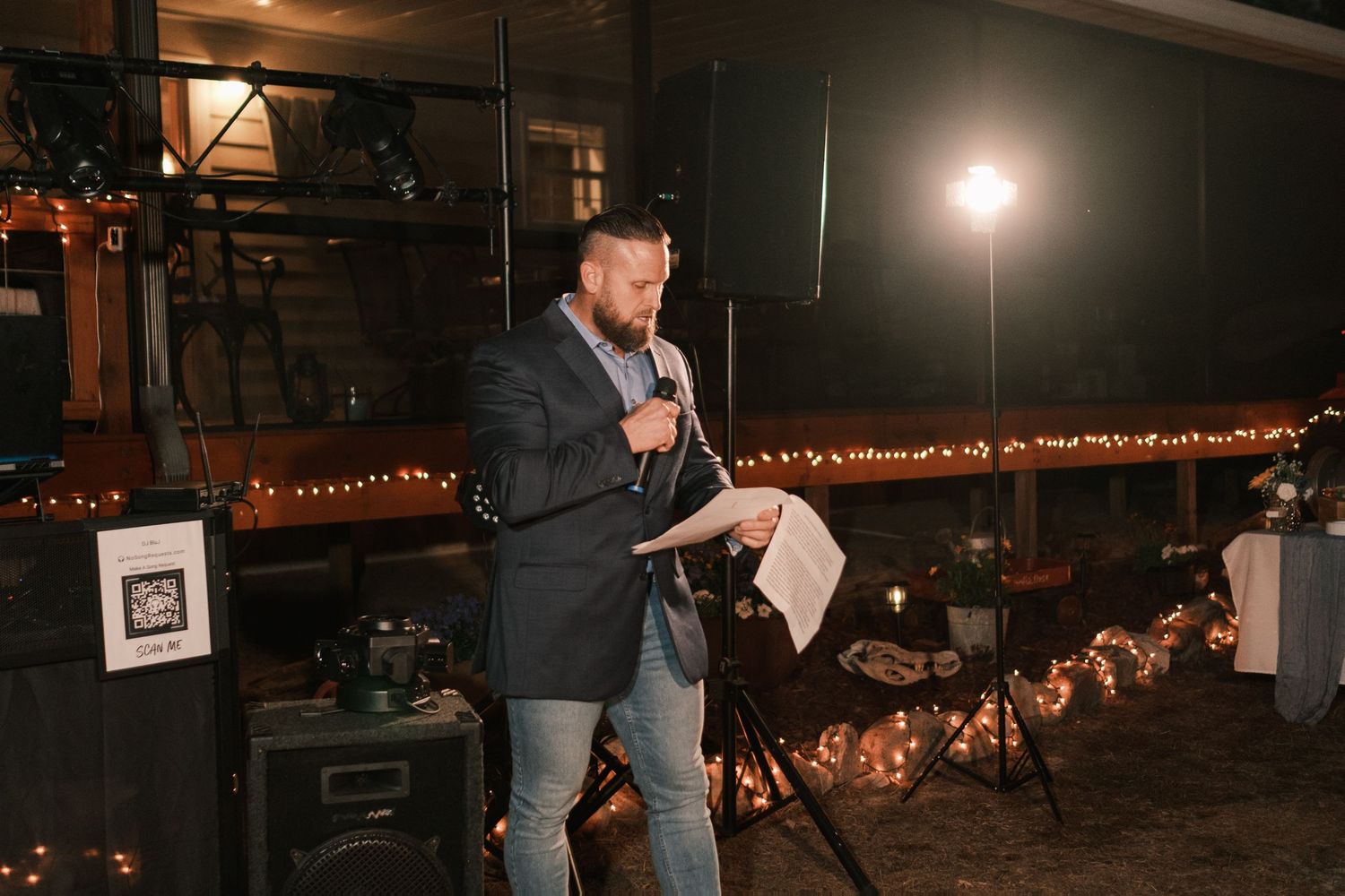 Wedding officiant or speaker reads from papers while holding a microphone at an outdoor evening reception.