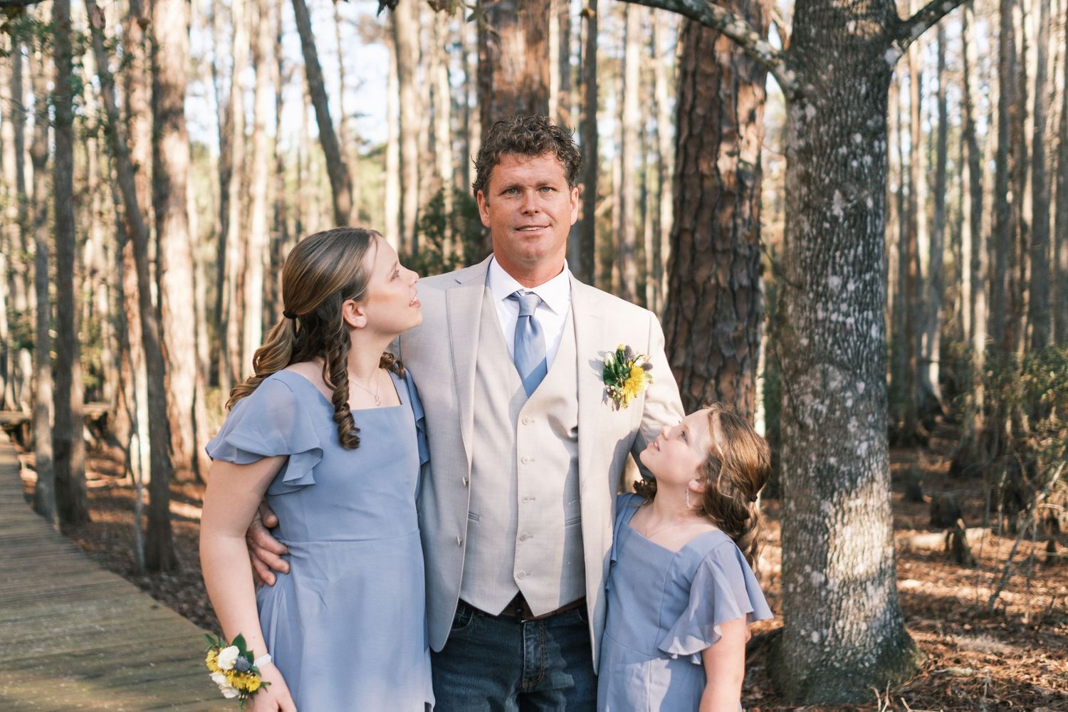 Groom poses with two young girls in blue dresses among tall trees at an outdoor wedding.