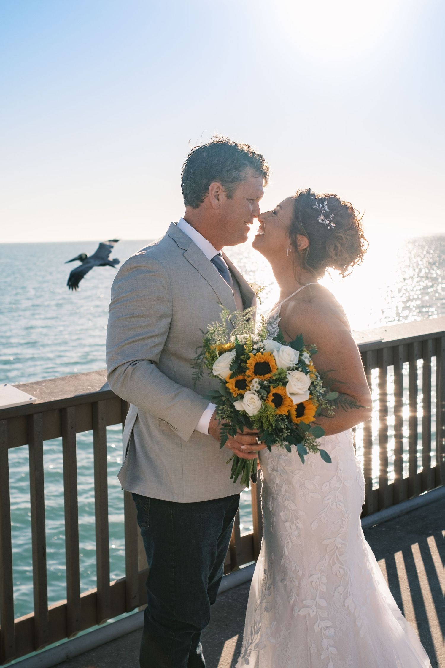 Bride and groom nearly kiss on pier at sunset, she holding sunflower bouquet, bird flying behind.
