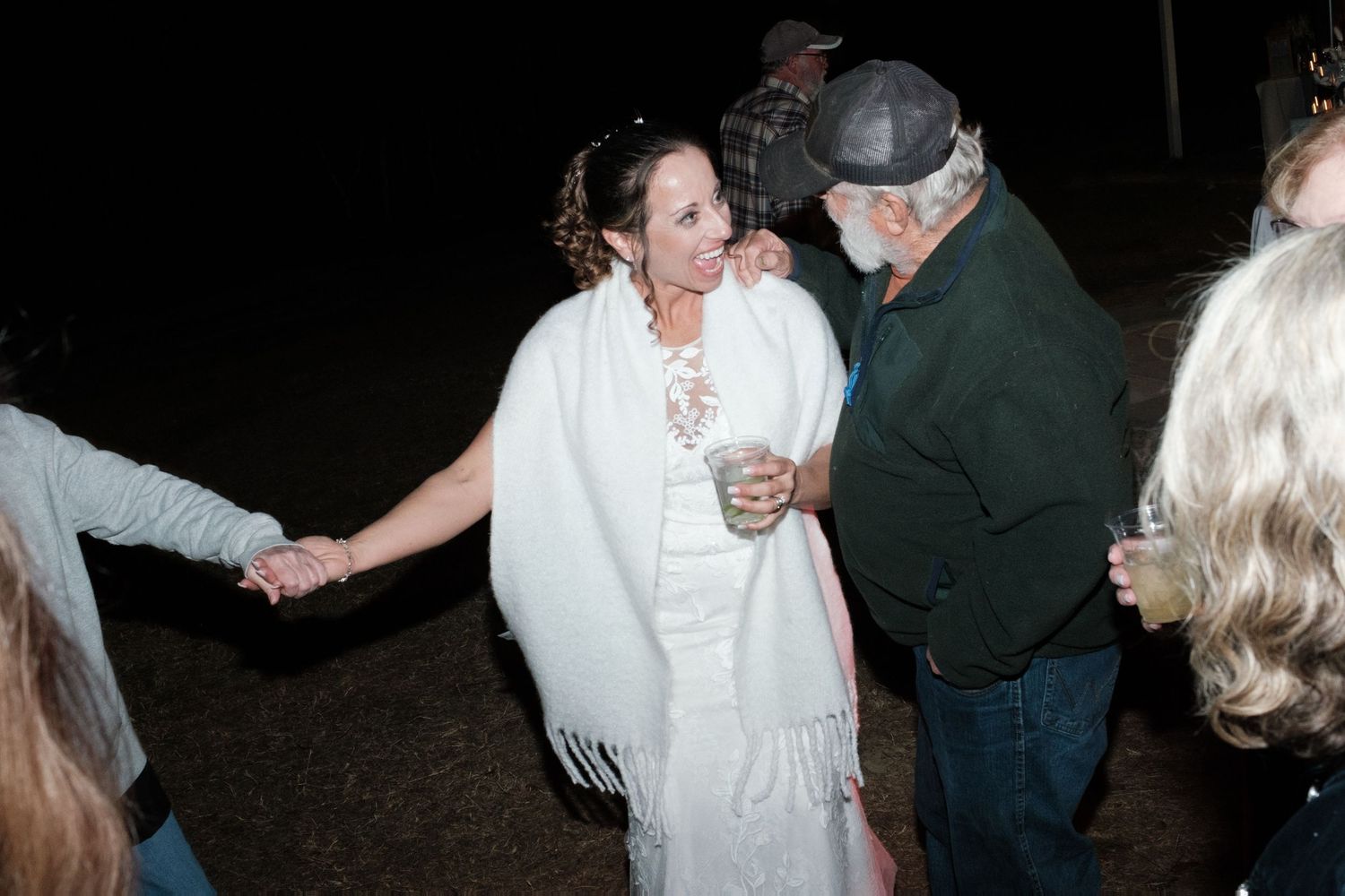 Bride in white wrap dancing and holding hands with an older guest at an outdoor nighttime wedding reception.