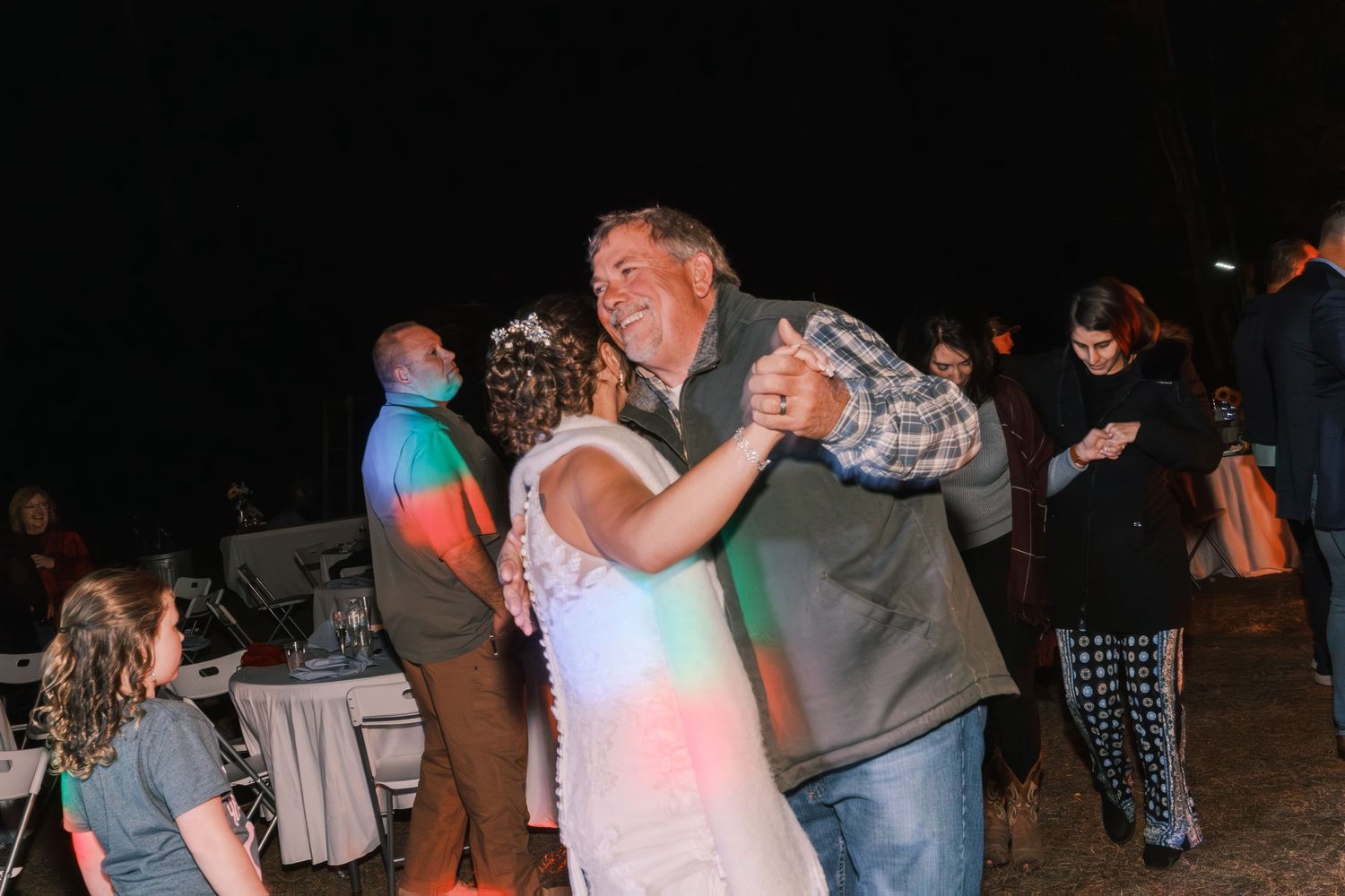 Bride dancing joyfully with older man in plaid shirt at outdoor evening wedding reception.
