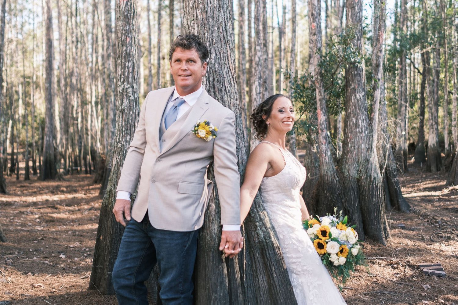 Bride and groom holding hands in pine forest, groom in tan suit with yellow boutonniere.