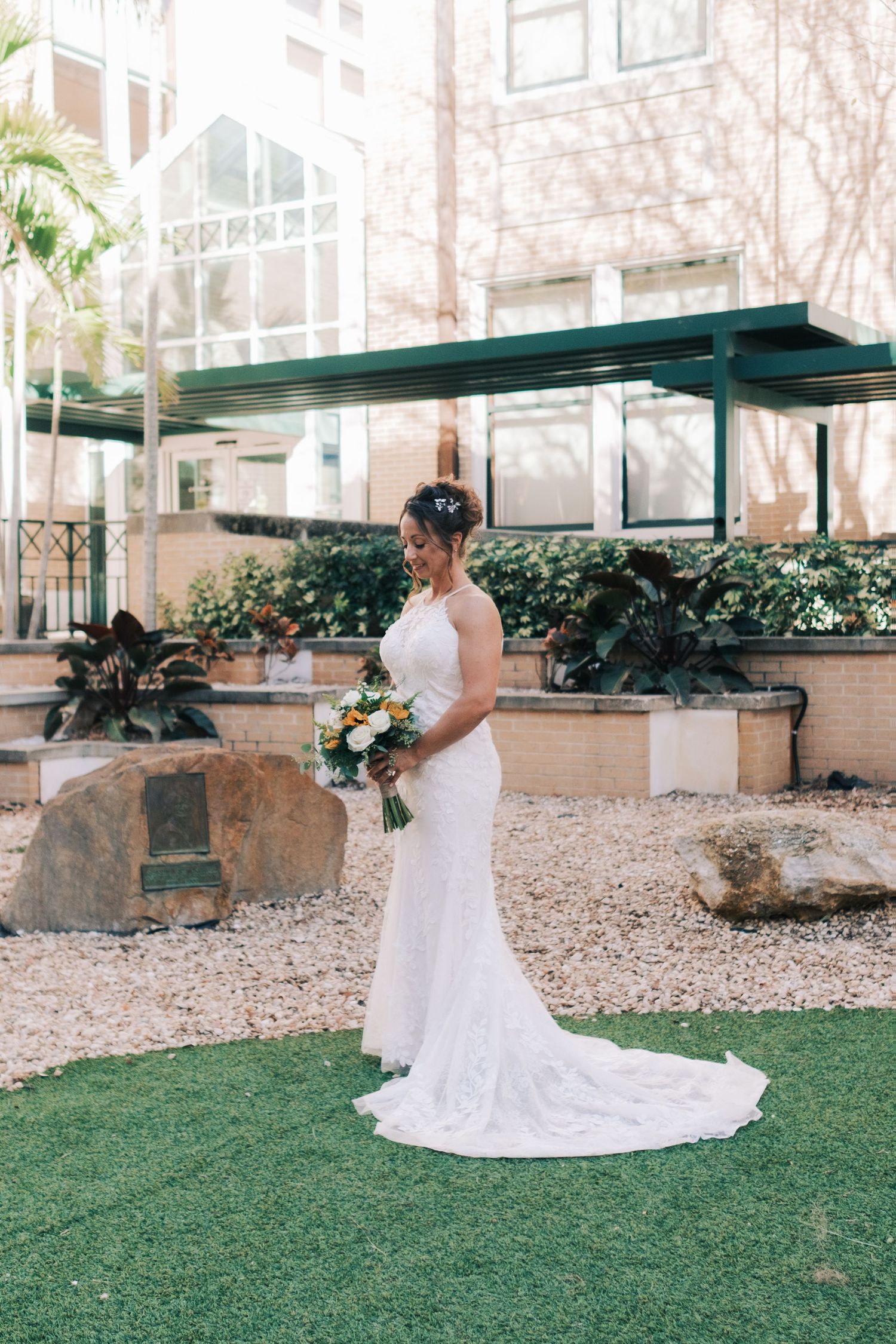 Bride in white lace mermaid gown holding bouquet standing in garden courtyard.