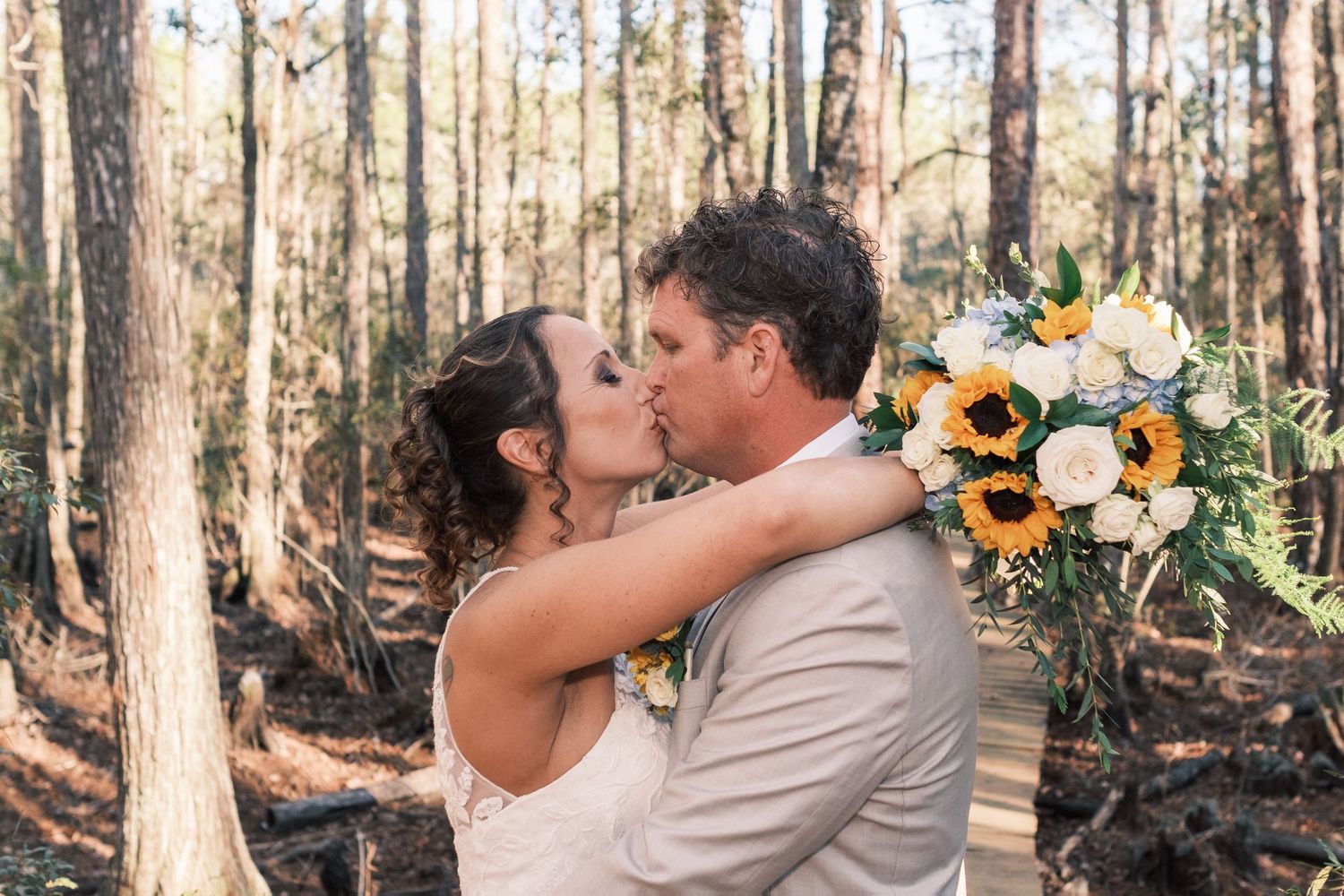 Bride and groom share a romantic kiss in a sunlit forest holding a sunflower bouquet.