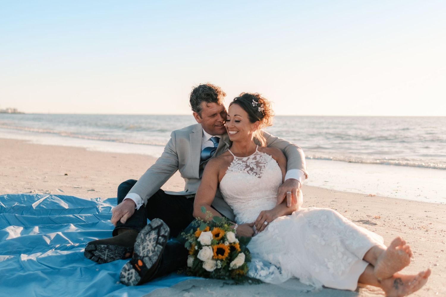 Bride and groom sit together on blue blanket on beach at sunset, laughing and holding bouquet.