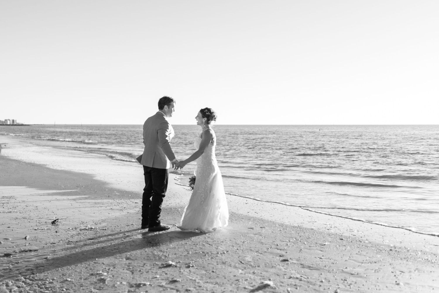 Black and white photo of bride and groom holding hands facing each other on serene beach.