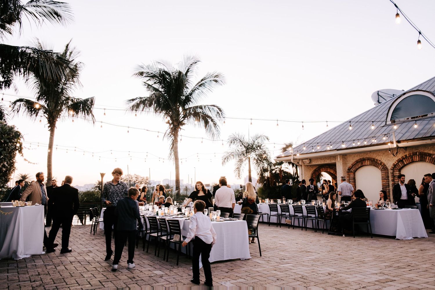 Outdoor evening wedding reception setup with string lights and palm trees at sunset.