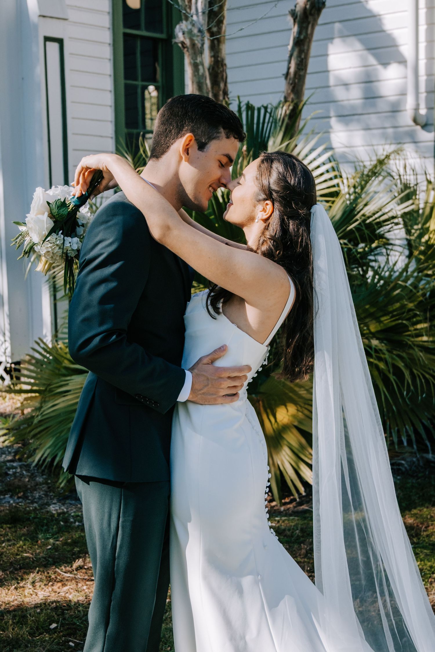 Intimate wedding couple portraits against white building exterior with flowing veil and palm trees.